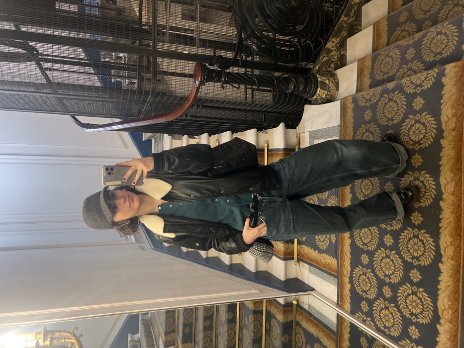 A young woman stands on a carpeted marble landing, taking a mirror selfie with a silver iPhone. She wears a dark gray cap, a black bomber jacket with a cream sherpa collar, a dark teal button-up cardigan, loose blue cuffed jeans, and black chunky loafers. Her right hand is in her jeans pocket, and she carries a small black-and-white patterned purse and a larger black shoulder bag.
The setting is a grand interior staircase of a building characteristic of Haussmannian architecture in Paris, France. The environment features white marble stairs with an ornate dark blue and gold floral-patterned carpet, light-colored walls with classical moldings, and a decorative gold chandelier partially reflected on the far left. To the right, an intricate black wrought-iron cage elevator shaft with a dark wooden handrail rises vertically. The woman stands directly on the patterned carpet, reflected in a large, unadorned mirror mounted opposite the elevator shaft.