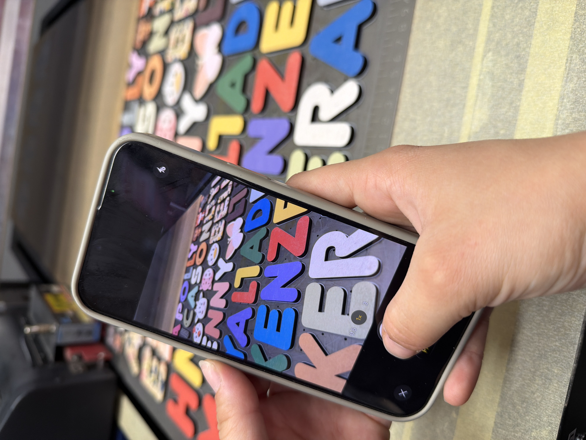 A close-up shot shows a person holding a smartphone and photographing colorful, raised alphabet letters laid out on a surface. The smartphone is held in a horizontal orientation, and the screen displays a camera interface with zoom controls. The letters are in various colors, including red, yellow, blue, green, purple, pink, orange, white, and brown, and appear to be made of a thick material, possibly foam or plastic. Some letters have small, cartoon-like images on them, such as a cat, a ladybug, and a butterfly. The background appears to be a dark, patterned surface, with other letters partially visible. To the left of the frame, a black and silver machine with red and yellow accents is partially visible, suggesting it may be involved in the production or arrangement of the letters. The scene appears to be indoors, likely in a workshop or creative space. There is no discernible information to place this scene in Ternopil, Ukraine.
