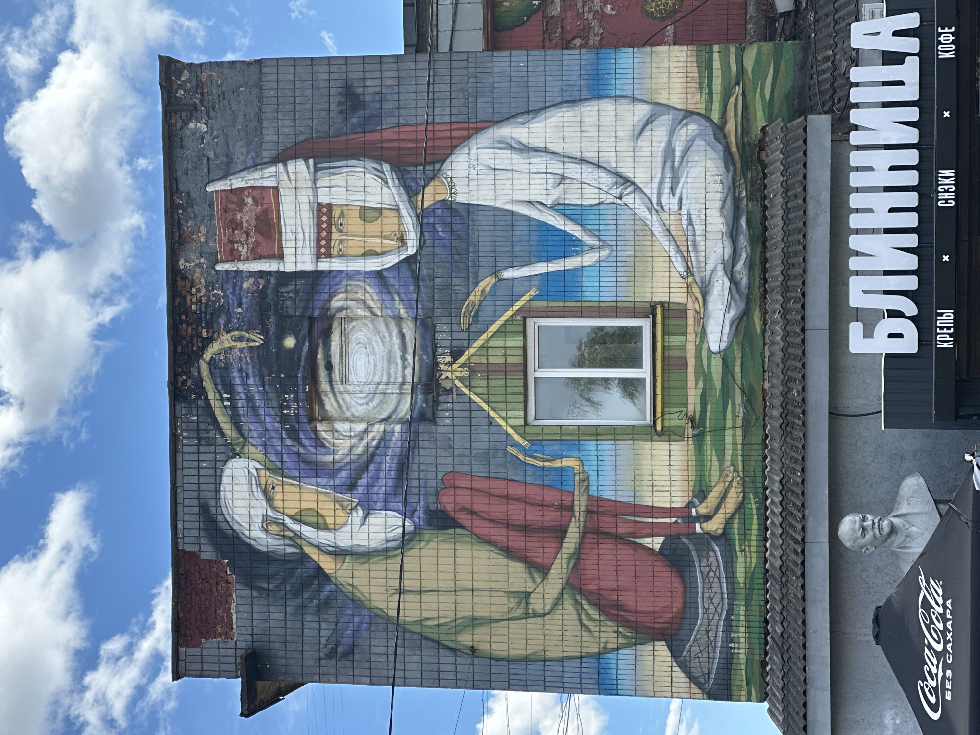 The image shows a mural on the side of a building in Minsk, Belarus. The mural depicts two figures with elongated limbs and stylized faces. One figure is wearing a white head covering with a red band. Both figures are holding long, yellow rods. The background includes swirling colors of purple, blue, and white. A window is incorporated into the mural's design. To the right of the mural is a sign in Cyrillic script that says "БЛИННИЦА". Below the sign is a sculpture of a man's head and an umbrella with "Coca-Cola" written on it in Cyrillic. The sky with white clouds is visible in the upper-left corner.