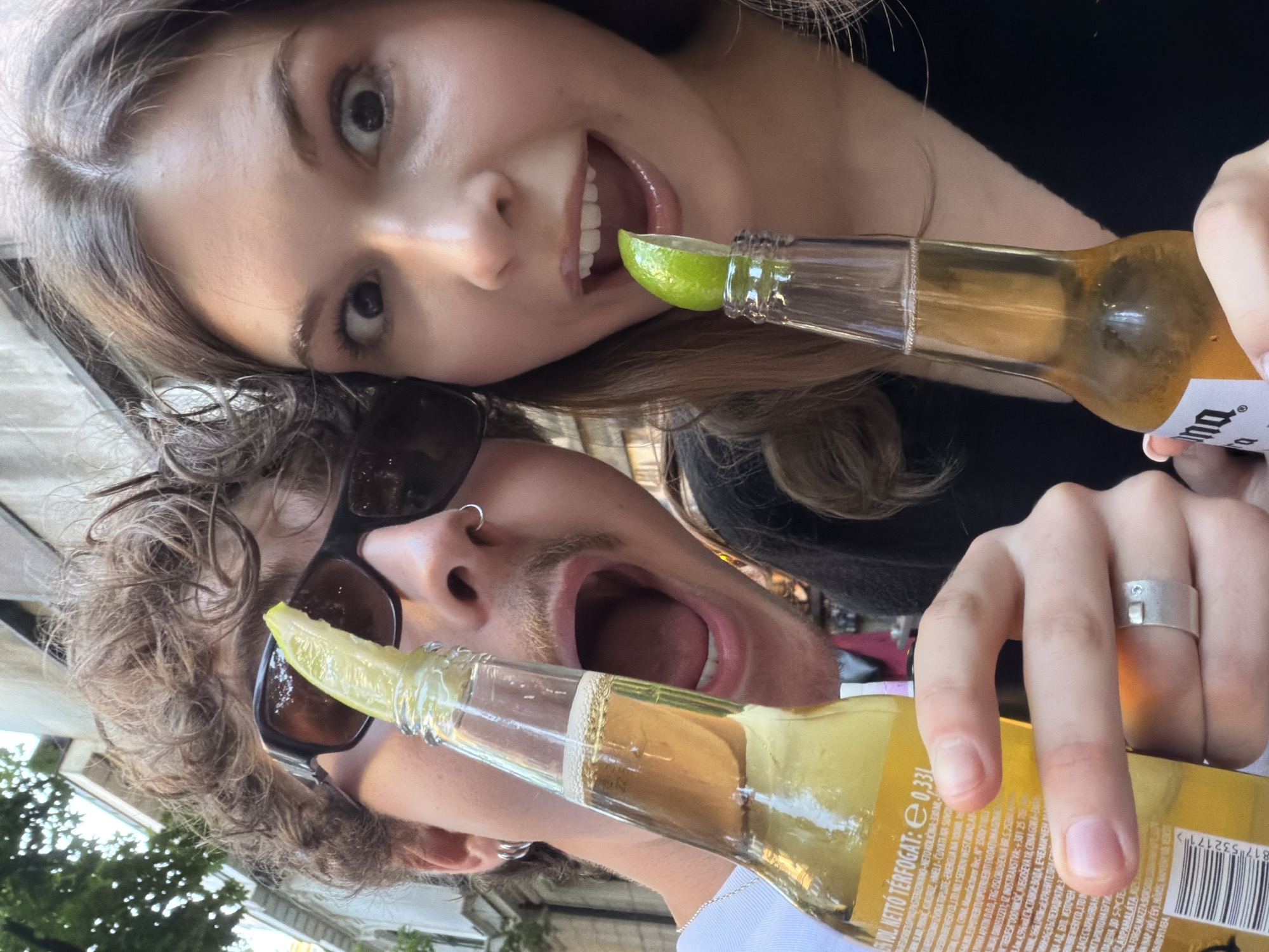 Two individuals, a male and a female, are posing for a selfie in an outdoor urban setting. The male, on the left, has curly brown hair, sunglasses, and a nose ring, and is holding a bottle of Corona beer with a lime wedge. The female, on the right, has long brown hair, and is also holding a bottle of Corona beer with a lime wedge. Both individuals have their mouths open and appear to be laughing or shouting. The background shows a building and trees, suggesting a street or a park in a city. Based on the text on the beer bottle, which includes "NETTO TÉRFOGAT: 0,33L," a Hungarian phrase meaning "NET CONTENT: 0.33L," and the style of the buildings, it is plausible that this image was taken in Belgrade, Serbia, a city with a significant Hungarian minority and a vibrant street life.