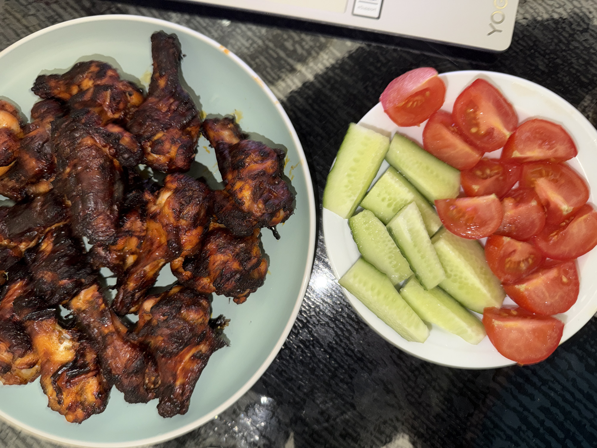 A close-up, top-down view shows two plates of food on a dark, textured surface. The upper plate, a light green ceramic, is filled with dark, glistening chicken wings, appearing to be roasted or grilled, with some parts slightly charred. Below this, a white plate holds chopped cucumbers and quartered tomatoes. The cucumbers are light green and appear fresh, while the tomatoes are bright red and juicy. To the right of the plates, a sliver of a silver laptop with the logo "YOG" is visible. The scene is lit by overhead lighting, casting subtle shadows. There are no people visible in the image. The environment appears to be an indoor setting, possibly a home or dining area, but the specific location in Bila Tserkva, Ukraine, cannot be determined from the visual information provided.