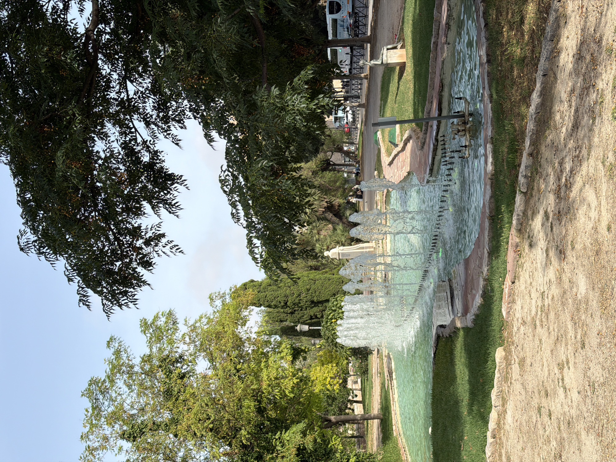 This media file shows a park scene with a fountain. The fountain is the central focus, with water spraying upwards in multiple jets and cascading into a pool. The pool has green water and is surrounded by grassy areas and a sandy path. In the background, there are trees, some with dark green foliage and others with lighter green leaves. Further back, there is a street with buses and some people walking. There are also some architectural elements visible, including a statue or monument and a lamppost. The sky is light blue with some overcast. The overall impression is a sunny day in a park. There are no visible signs of text in this media file. The time of day appears to be daytime, and the weather seems pleasant.