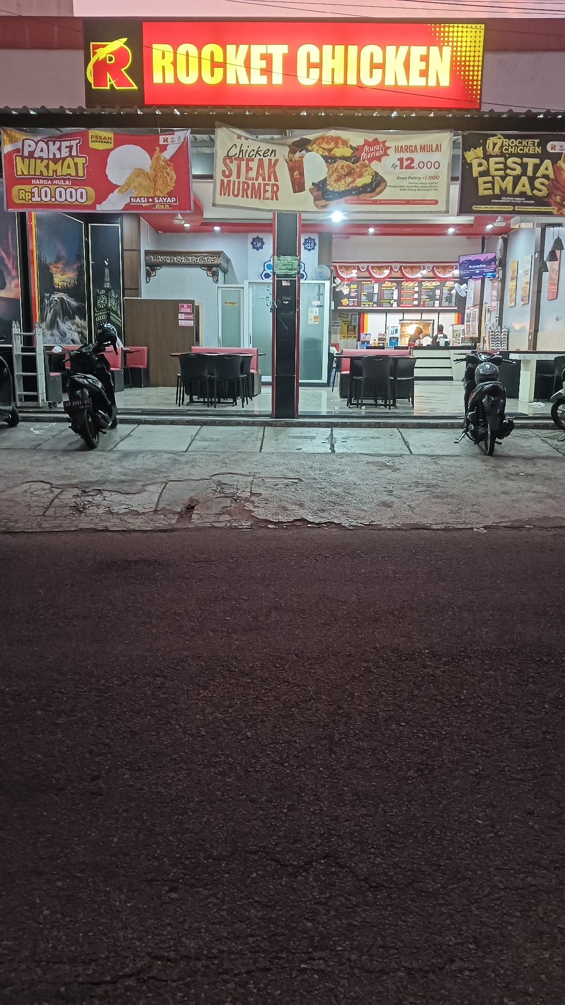 The image shows the exterior of a Rocket Chicken restaurant in Sragen Tengah, Indonesia. A bright red sign with yellow lettering displays "ROCKET CHICKEN". Below the sign, there are several banners advertising food items and prices. One banner shows "PAKET NIKMAT Rp.10.000" with an image of fried chicken and rice. Another banner reads "Chicken STEAK MURMER HARGA MULAI Rp.12.000". A third banner is for "ROCKET CHICKEN PESTA EMAS".
The restaurant has a glass storefront revealing the interior, which includes tables and chairs. There is a counter area with menu displays and staff members visible in the background. Outside the restaurant, two motorcycles are parked on the sidewalk. The overall scene depicts a commercial establishment at night, illuminated by the restaurant's signage and interior lights. The foreground shows a paved road with some cracking.