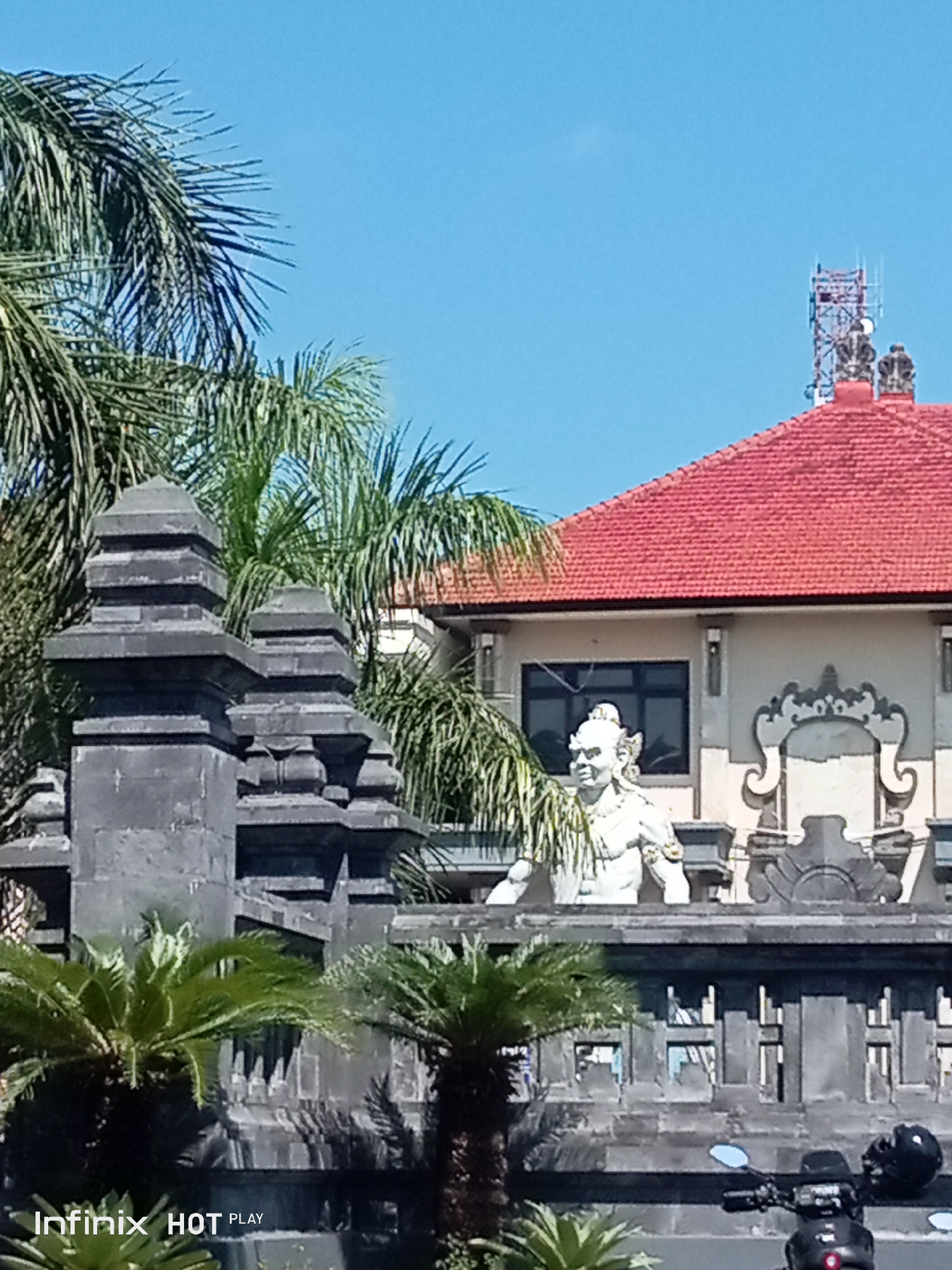The image shows a statue of a white mythical figure in front of a building with a red tiled roof. Palm trees are visible on the left and in the background. There is also a cell tower on the roof of the building. In the foreground, there is a stone wall and a motorcycle. The bottom left corner of the image shows the text "Infinix HOT PLAY". The scene appears to be in Gianyar, Indonesia, judging by the architectural style and vegetation.