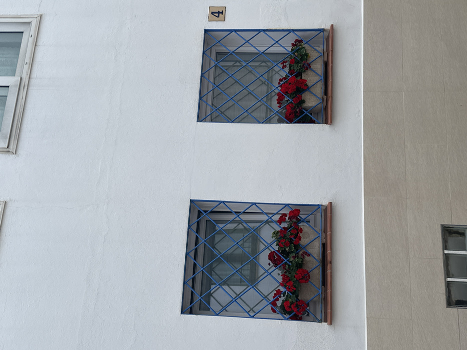 A portion of a building facade in Calella De Palafrugell, Spain, is depicted. The upper section of the facade consists of white textured stucco, while the lower section features light beige rectangular tiles. Two identical, square windows are visible in the middle section, each fitted with a blue metal security grille forming a diagonal grid pattern. Below each window, a terracotta-colored sill supports a rectangular, light-colored planter box. Both planter boxes contain vibrant red geraniums and green foliage. A small, square ceramic tile bearing the black numeral '4' is affixed to the white stucco wall to the right of the rightmost window. Partial views of additional windows are present at the top and bottom edges of the frame. No individuals or discernible actions are depicted in the scene.