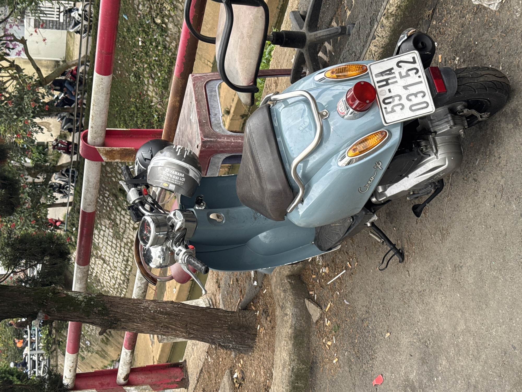 The image shows a light blue scooter parked on a paved surface next to a tree. The scooter has a dark brown seat and a license plate that reads "59-HA 031.52". Two helmets are resting on the handlebars. In the background, a red and white striped railing runs along a canal. Beyond the railing, there are more trees and several other motorcycles are parked, with a few people visible in the distance. The overall scene suggests a street scene in Da Lat, Vietnam, characterized by the presence of motorcycles, the style of architecture in the background, and the vegetation. The ground around the scooter is littered with cigarette butts.