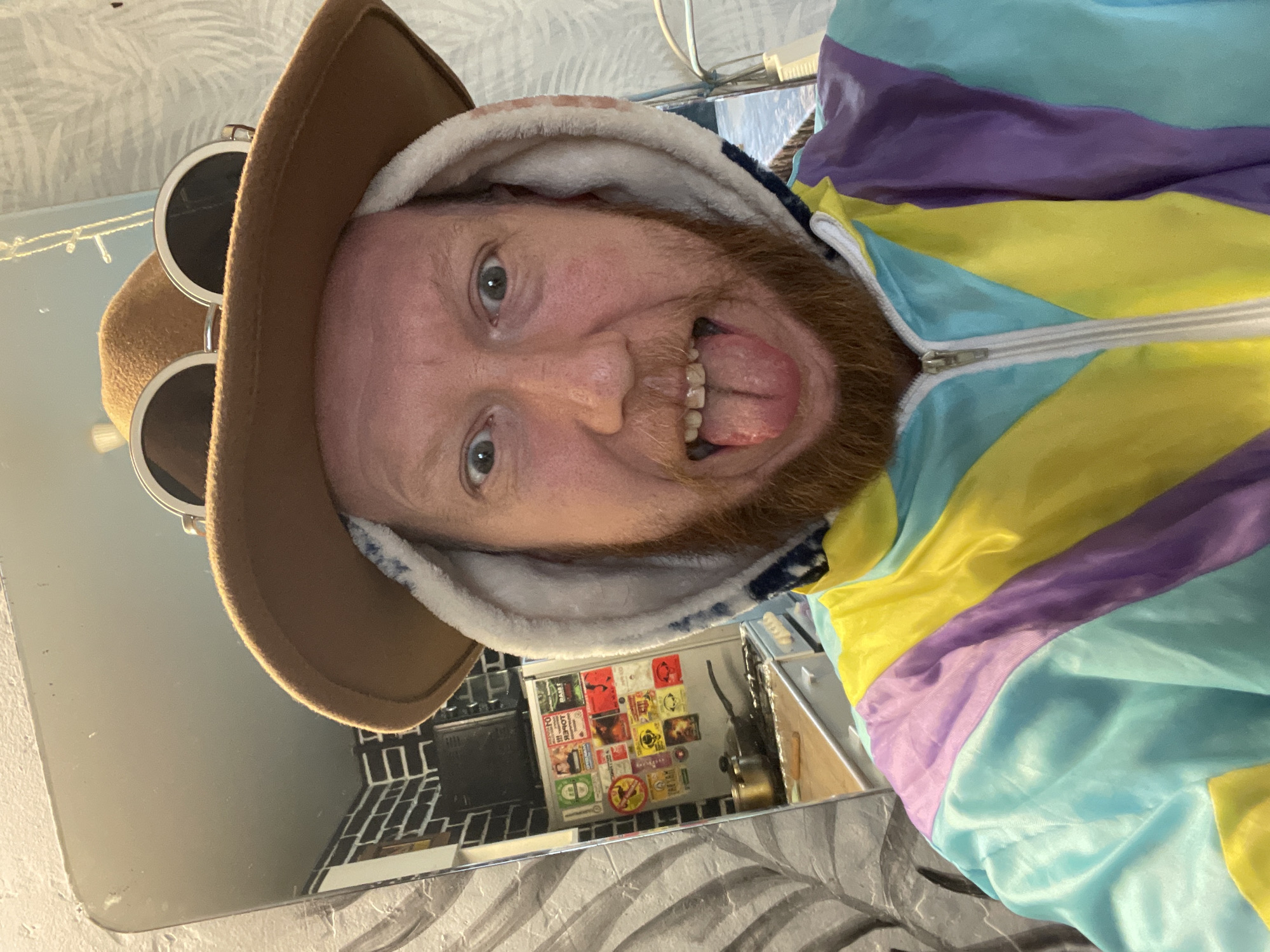 A man with a beard and ginger hair is taking a selfie in a bathroom. He is wearing a brown cowboy hat with round sunglasses perched on top, a hooded jacket with white trim, and a colorful windbreaker with pastel stripes of yellow, purple, and turquoise. His tongue is sticking out and his eyes are wide open. He is reflected in a mirror to his left, which shows a kitchen with a refrigerator covered in stickers and a stovetop with a pot. The background of the bathroom appears to be a wall with a textured, gray and white pattern. The overall scene suggests a playful and informal setting.