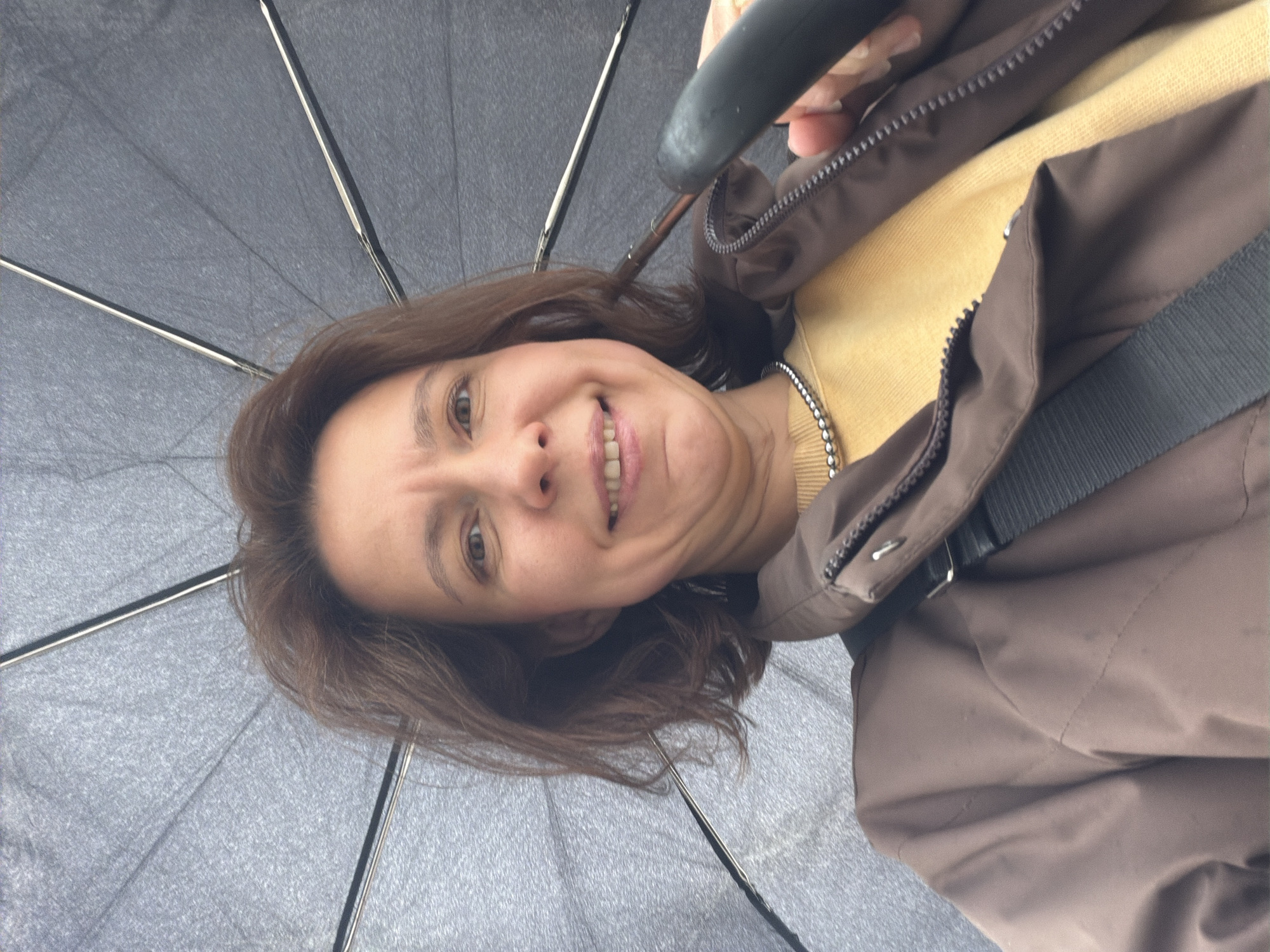 A woman is smiling at the camera while holding an open umbrella. She is wearing a yellow shirt and a brown jacket. The umbrella is gray with silver ribs. The location is unknown, and there is no information to suggest it is Chaikovsky, Russia.