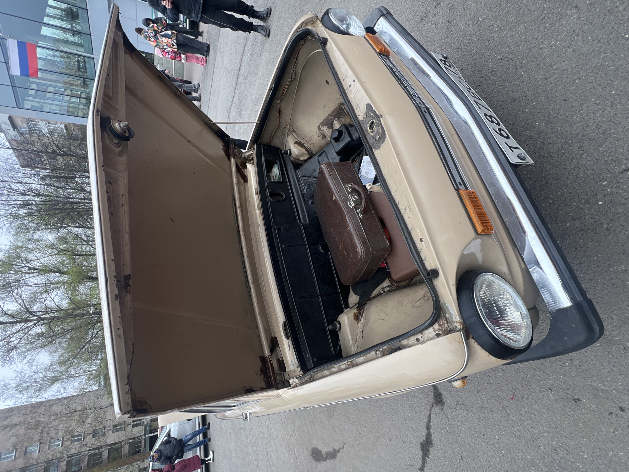 The image captures the front of a vintage beige car with its hood open, revealing a brown suitcase and a black box in the storage area. The car has a rectangular license plate that reads "T687BC 78," indicating it is in Russia. The surrounding environment suggests an urban setting with a building in the background, identifiable by the presence of a Russian flag. There are people in the background, who seem to be observing the car. The overall scene suggests that the photograph was taken in Saint Petersburg, Russia.