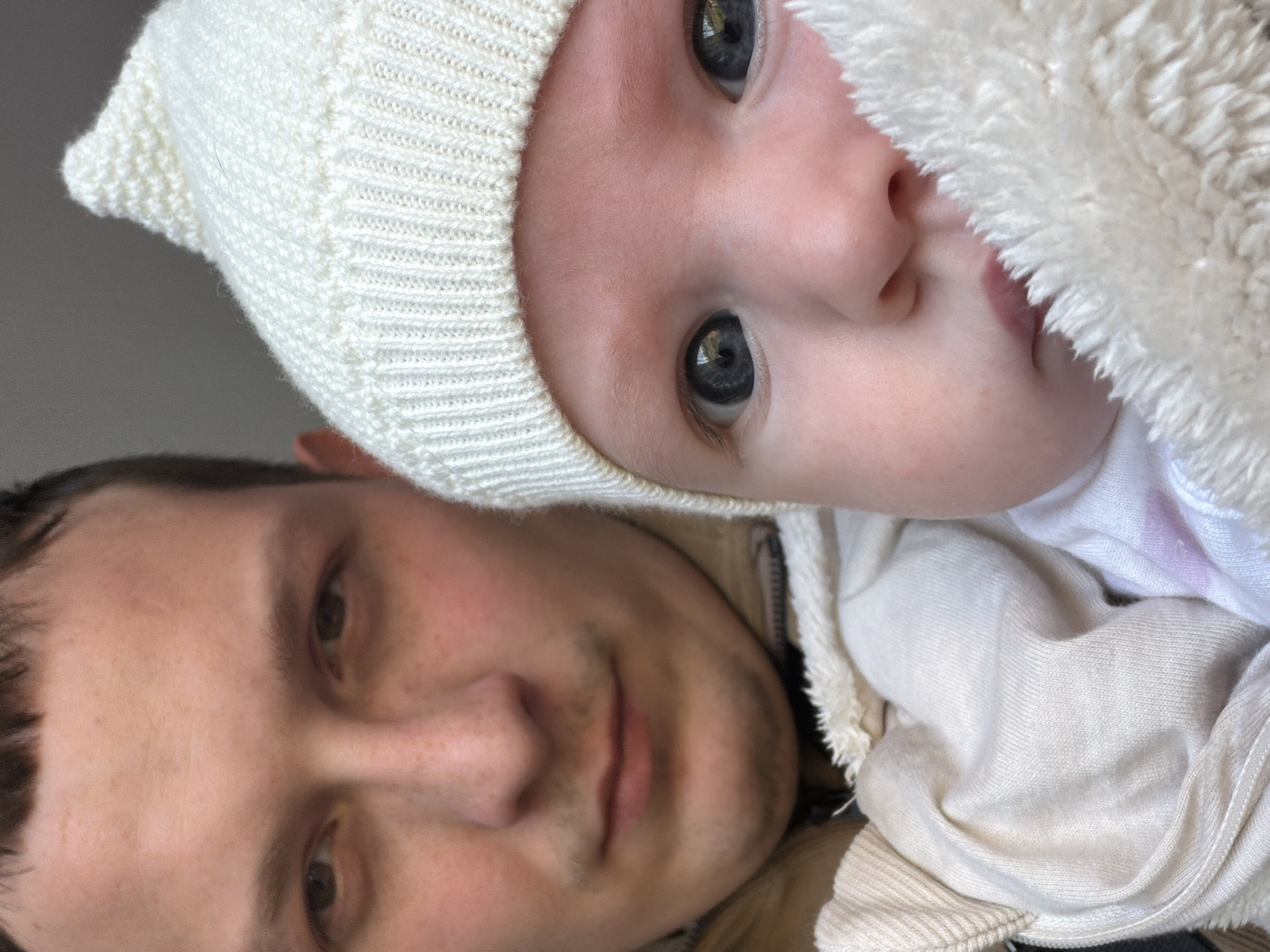 A close-up shot shows a man and a baby. The baby, wearing a white knit cap, is positioned in the upper portion of the frame. A fluffy, white blanket covers part of the baby's face. The man, presumably holding the baby, occupies the lower half of the image. The background appears to be a neutral color. There is no information to suggest the image was taken in Chaikovsky, Russia.
