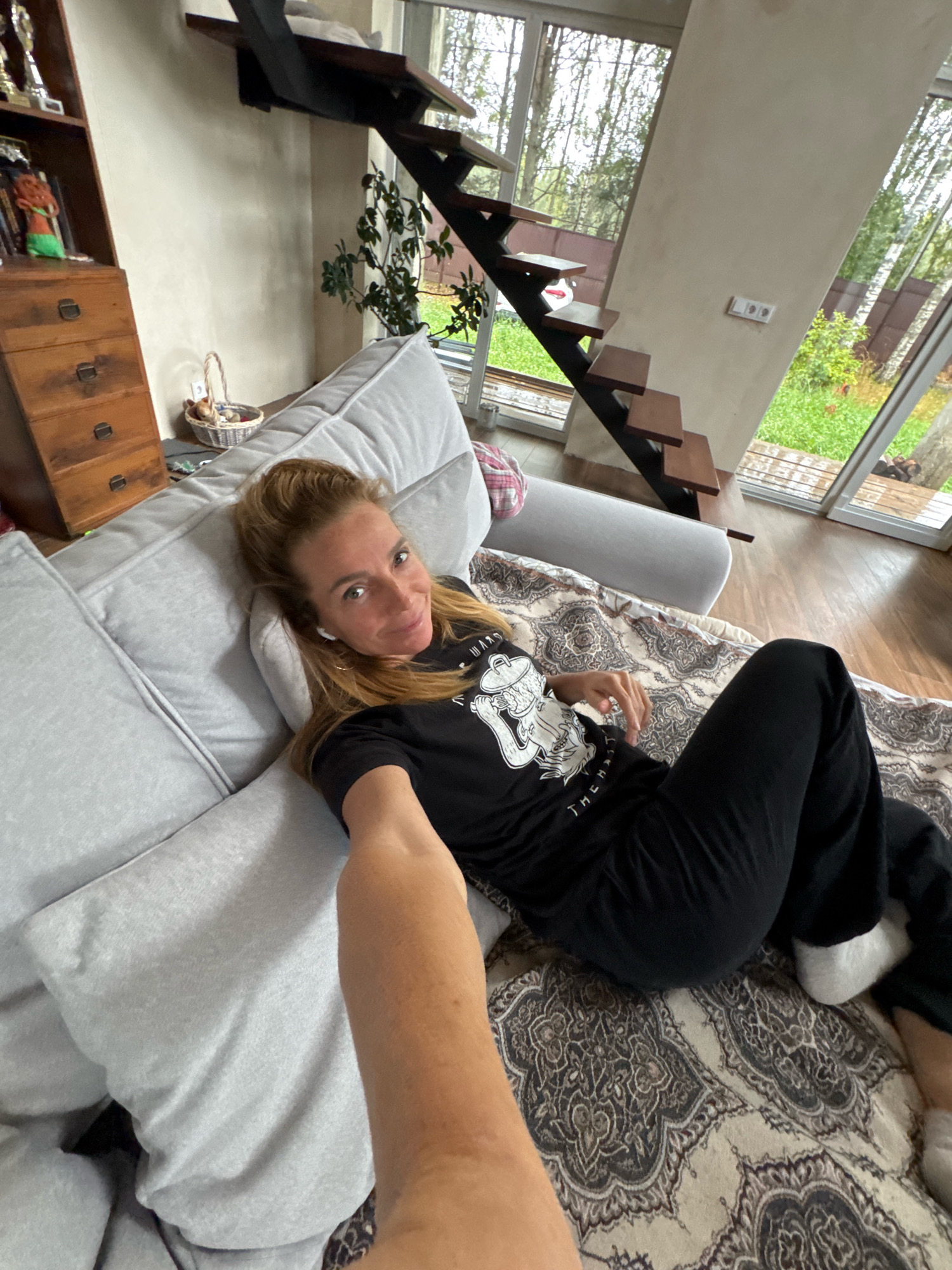 An indoor, eye-level, medium shot captures a blonde woman lying on a light gray sofa in what appears to be a residential living room. She is wearing a black t-shirt with a graphic design and black pants. Her left arm is extended towards the camera, holding the device used to take the photo. She is wearing white wireless earbuds. The sofa is adorned with a patterned blanket.
Behind the sofa, a wooden dresser with four drawers is visible. To the left of the dresser, a shelf holds several items, including trophies and decorative objects. A small wicker basket with some contents sits on the floor between the dresser and the sofa.
In the background, a modern staircase with wooden steps and black metal railings ascends to an upper level. Large windows with a view of trees and greenery suggest an outdoor environment. The room has light-colored walls and wooden flooring. The overall impression is of a casual, comfortable indoor setting.