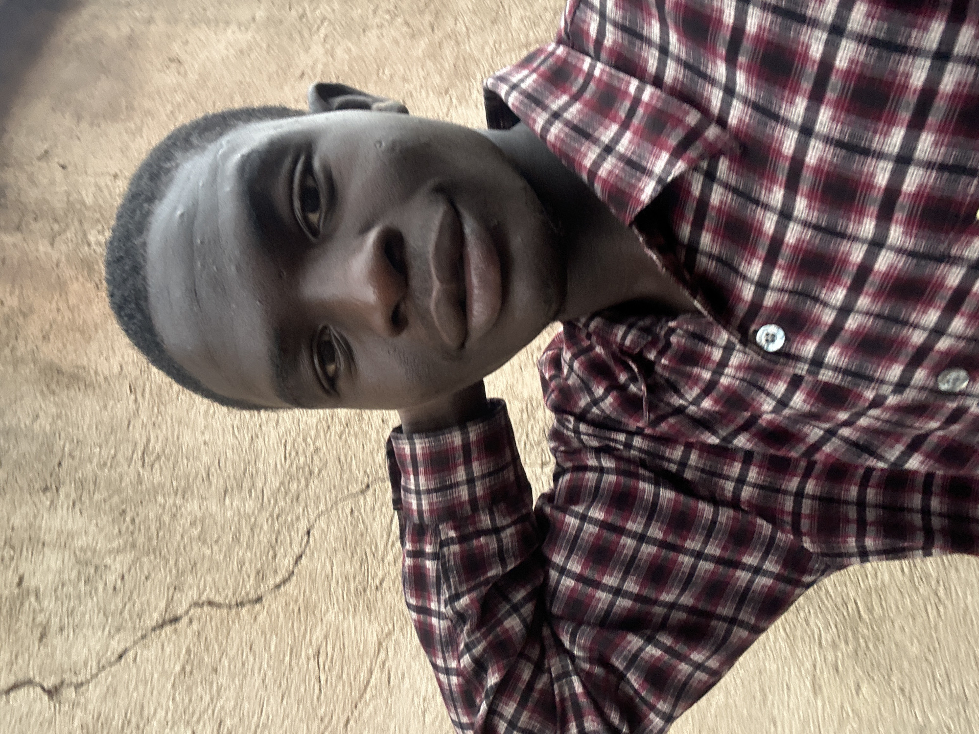This media file features a close-up, rotated portrait of a young Black person wearing a maroon, white, and black plaid button-up shirt. The individual's head is tilted, and their left arm is bent, supporting their head, with their hand resting on their forehead. Their gaze is directed towards the viewer, and they have a neutral to slightly curious expression. The background consists of a textured, light brown surface, possibly a wall or ground, with visible cracks. The lighting suggests it is daytime, and the overall tone is casual. The setting appears to be outdoors, given the natural lighting and textured background, possibly in Jos, Nigeria. No text is visible in the image.