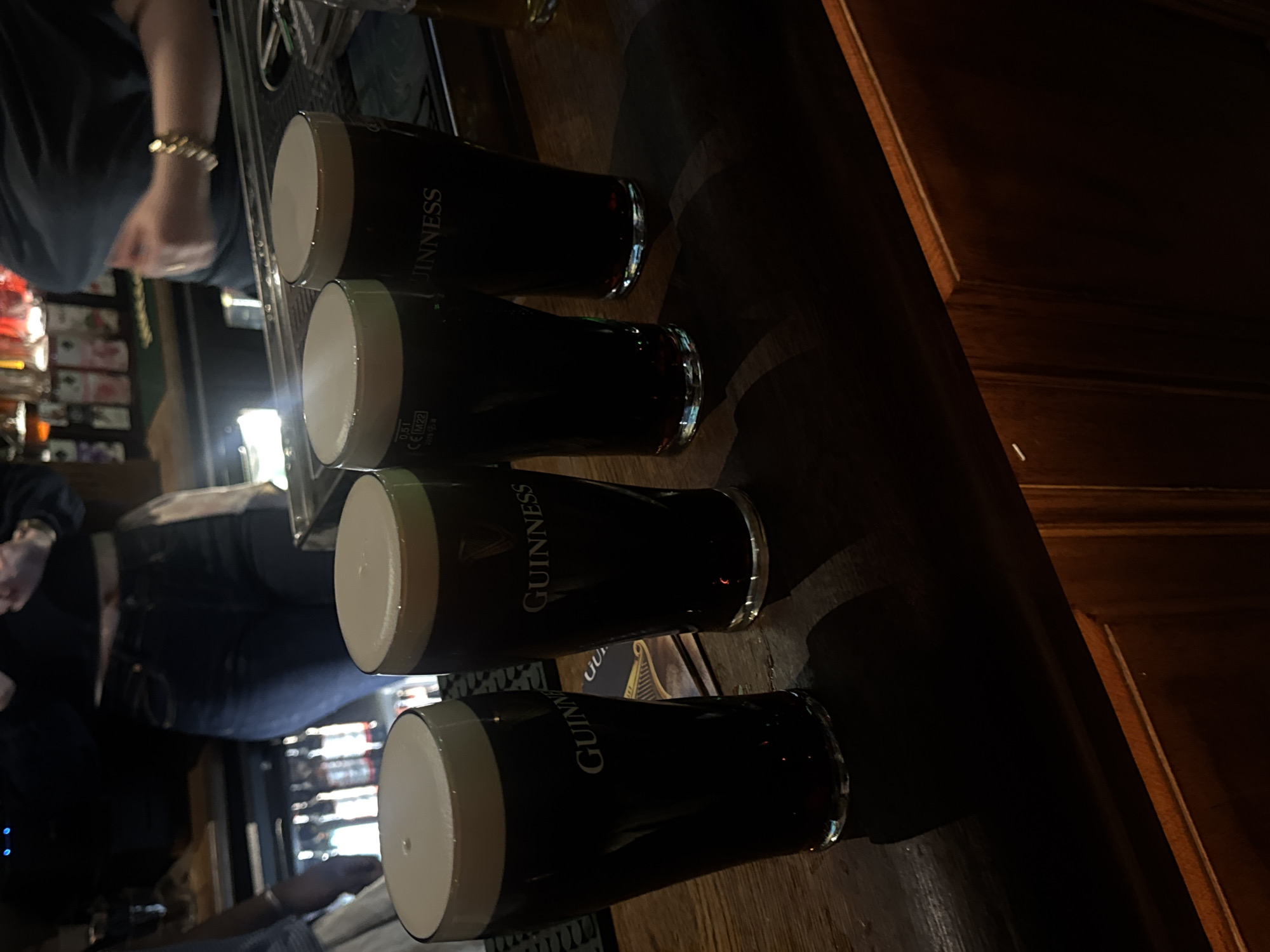 The image shows four pints of Guinness lined up on a wooden bar top. A person wearing jeans is partly visible to the left of the frame. The lighting is dim, and the environment appears to be a bar or pub. The reflection from glass and stainless steel lines the back wall to the left. "Guinness" is printed on the glasses. The location is indeterminable and cannot be definitively stated as being Paris, France.