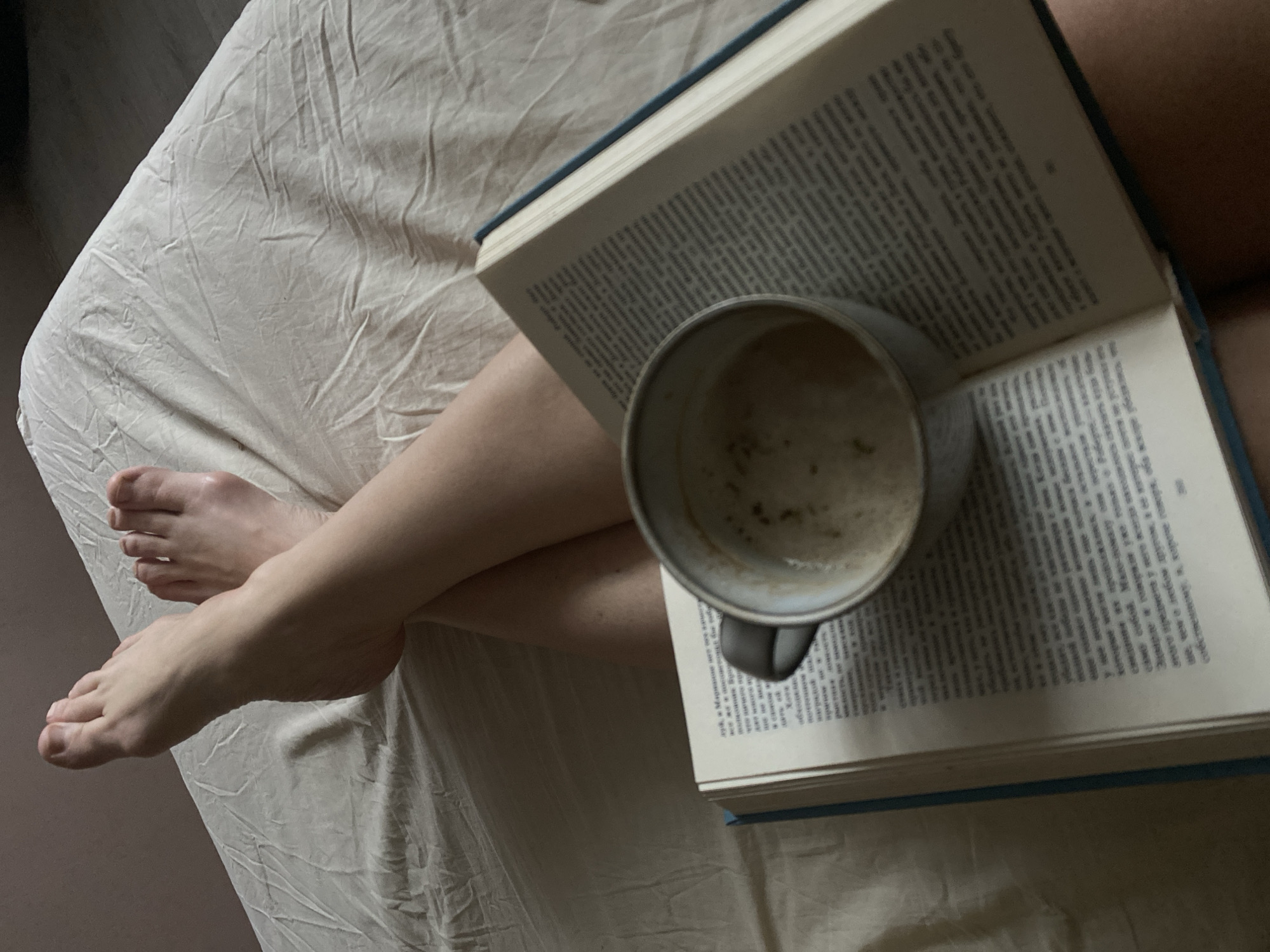 The image depicts a person lying on a bed with their legs crossed. An open book rests on their legs, and a partially consumed beverage in a mug sits on top of the book. The setting appears to be indoors, likely in a bedroom or similar space, given the presence of the bed. No information indicates the scene's location in the city of Chaikovsky, Russia.