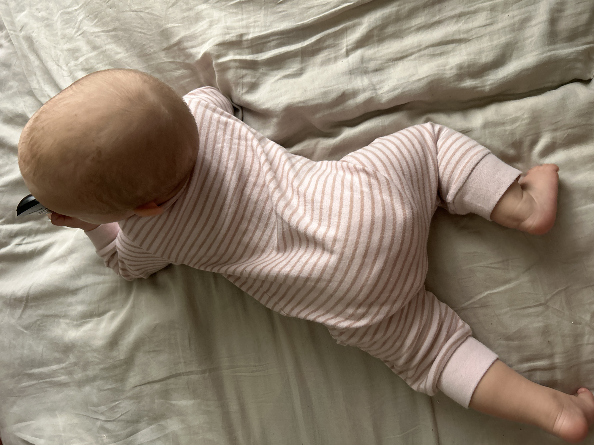 The image shows a baby lying on its stomach on a light-colored, wrinkled fabric surface, likely a bed sheet or blanket. The baby is viewed from above, mostly from the back, revealing the crown of its head with fine, light-colored hair. It is dressed in a full-body onesie featuring thin, vertical stripes in light pink and a very light beige or off-white. The baby's arms appear to be tucked underneath its chest, and its legs are slightly bent, with bare feet visible towards the lower right of the frame. The surface the baby is on is uniform in color, with many folds and creases. No specific activity beyond resting or being in a prone position is clearly discernible. The scene appears to be indoors, possibly during the day, given the soft, even lighting. No emotions are visible as the baby's face is obscured. No text is present in the media file. While the location context is Moscow, Russia, there are no specific visual cues within the image to indicate this.