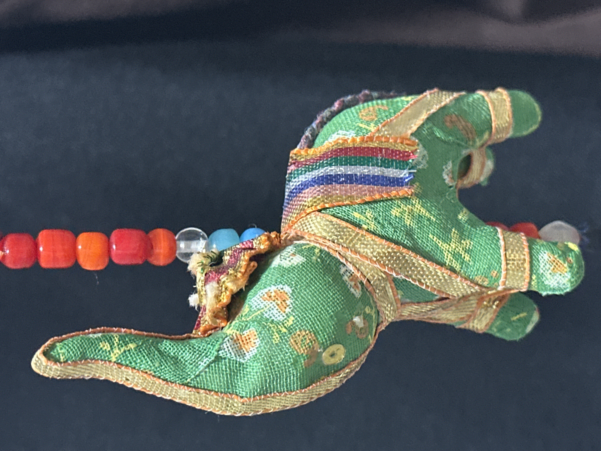 An overhead shot shows a green fabric elephant ornament with gold trim, a colorful striped blanket, and a beaded string above it. The ornament is hanging against a dark, blurred background. The elephant's trunk is curled upwards and its ears are large and pointed. The blanket on its back is made of vertical stripes of red, orange, yellow, green, blue, and purple. The beaded string above the elephant is composed of several red, orange, and blue beads, with one clear bead. The elephant appears to be a decorative item, possibly a keychain or a charm. The image does not contain any information about the city of Nevinnomyssk, Russia.