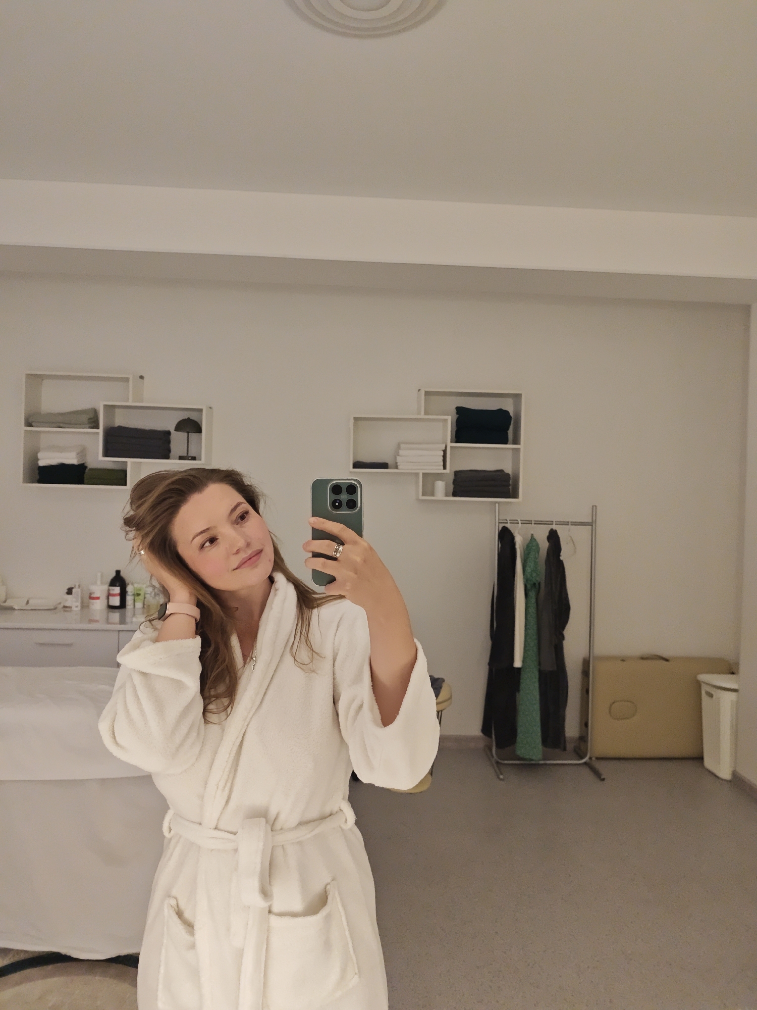 A woman is taking a mirror selfie in a white bathrobe in what appears to be a spa or salon. She is standing in front of a mirror, holding a smartphone in her right hand, and has her left hand on her hair. The room is clean and well-lit, with white walls and shelves displaying folded towels and some toiletries. There is a massage table covered with a white sheet on the left side of the frame, and a clothing rack with several garments on the right. A folded massage table and a trash can are also visible in the background. The overall impression is of a professional and relaxing environment. The city of Vinnytsia, Ukraine, is not identifiable from the visual cues in this image.