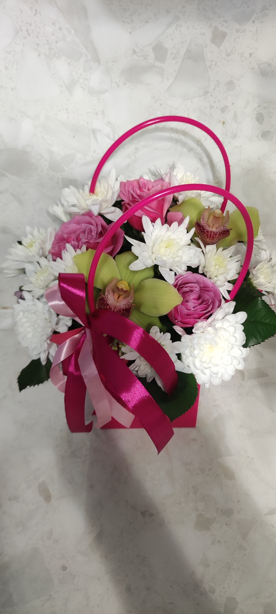 A bouquet of flowers is positioned in a bright pink container with matching handles. The flowers include white chrysanthemums, pink roses, and green orchids. A pink ribbon is tied in a bow around the container. The background appears to be a white, speckled surface. The location and its relation to Chaikovsky, Russia, are unknown.
