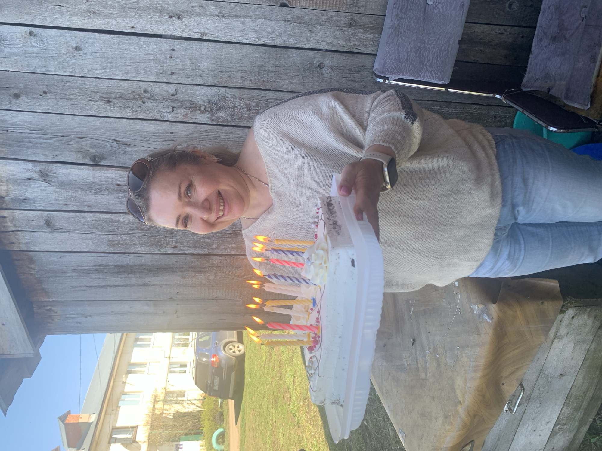 A woman holds a cake with lit candles. She wears sunglasses and a light-colored sweater with jeans. The background consists of a wooden fence and a street scene with buildings and a parked car. The photo was taken in the city of Кемуль, Russia.