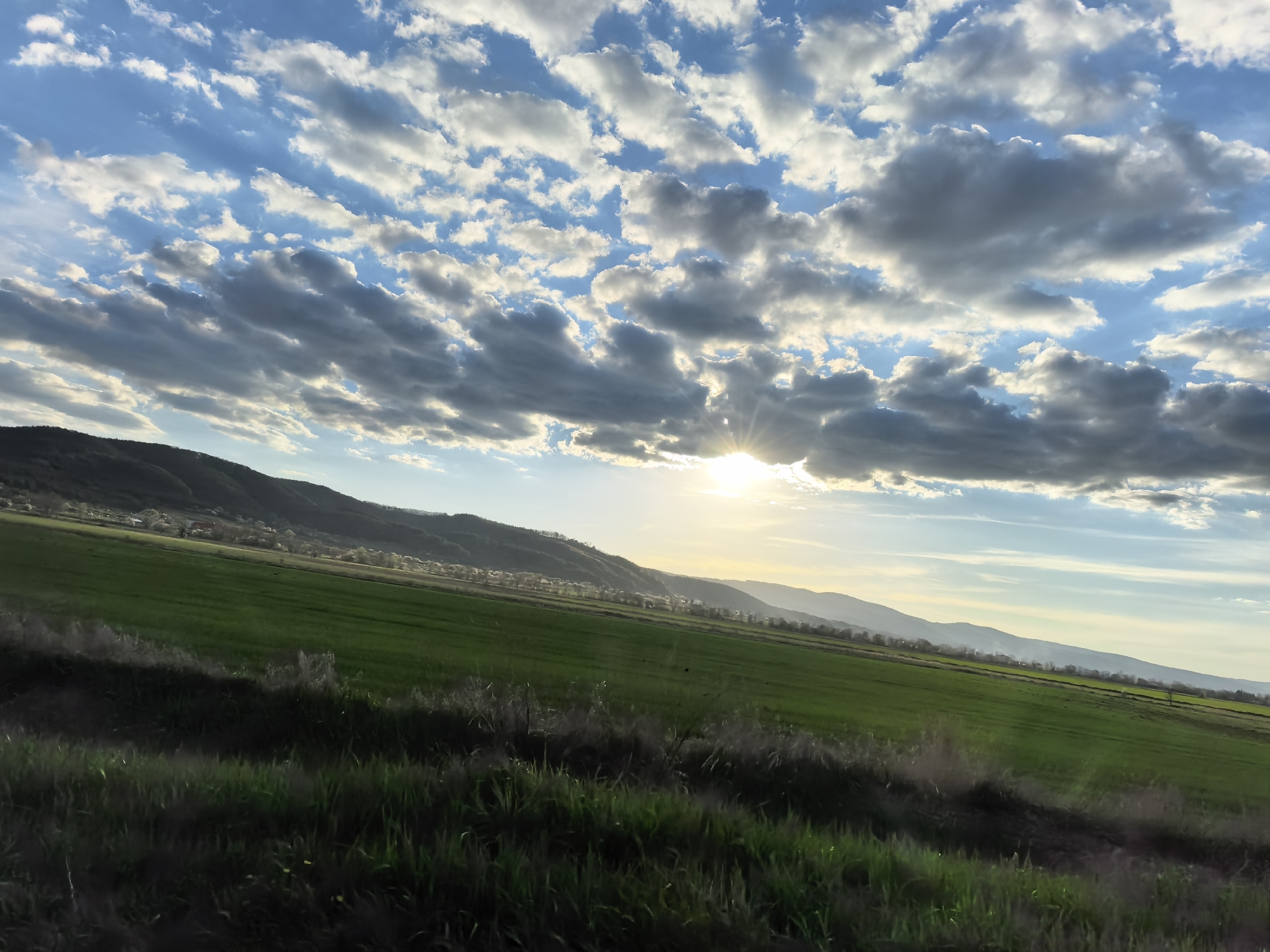 A wide-angle landscape photograph captures a serene rural scene, likely during sunrise or sunset, characterized by a dynamic sky, rolling hills, and lush green fields.
The upper half of the image is dominated by a partially cloudy sky. Patches of clear blue sky are visible, interspersed with numerous cumulus and stratocumulus clouds. These clouds vary in density and brightness, with some appearing brilliant white and luminous where the sun's rays hit them, while others are darker grey in shadow. A prominent sunburst is visible towards the upper right-middle, piercing through a gap in the clouds and casting bright, golden light across the scene.
Below the sky, a line of undulating hills or mountains stretches across the horizon from the left to the right. The hills on the left appear closer and are covered in darker green vegetation, with faint hints of structures or a small village nestled along their base. Towards the right, the distant mountains appear lighter and hazier, fading into a soft blue hue, indicative of atmospheric perspective.
In the midground, vast expanses of vibrant green fields slope gently down from the base of the hills. These fields appear to be cultivated land, possibly crops, and are illuminated by the soft, warm light of the low sun.
The foreground consists of wilder, darker green grass and vegetation, which appears to be a verge or roadside area. This section is darker, suggesting it is either in shadow or less directly illuminated by the sun.
There are no discernible people, animals, or artificial objects other than the potential distant village. No visible text is present in the image. The overall impression is one of natural beauty and tranquility.