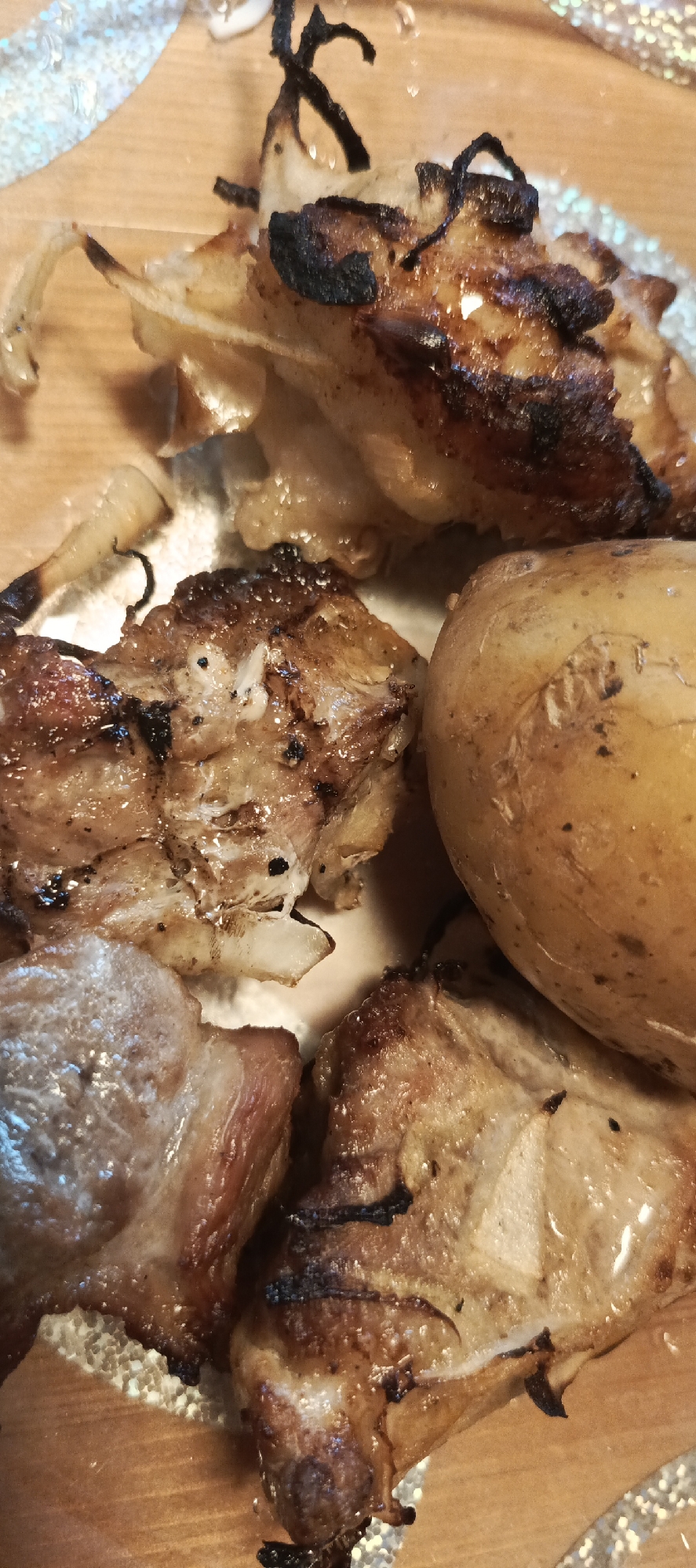 A close-up, overhead shot shows a portion of a meal on a wooden surface, likely a table. The meal consists of several pieces of roasted meat, appearing to be pork or chicken, with some charred edges and visible fat. There is also a whole roasted potato, golden brown in color, and some pieces of cooked onion, also showing signs of charring. The lighting is warm, highlighting the textures and colors of the food. The background is blurred, but a shimmering, textured edge, possibly from a decorative placemat or napkin, is visible on the left and right sides of the frame. The scene is located in the SNT (garden non-profit partnership) of Merchalovo, Russia.