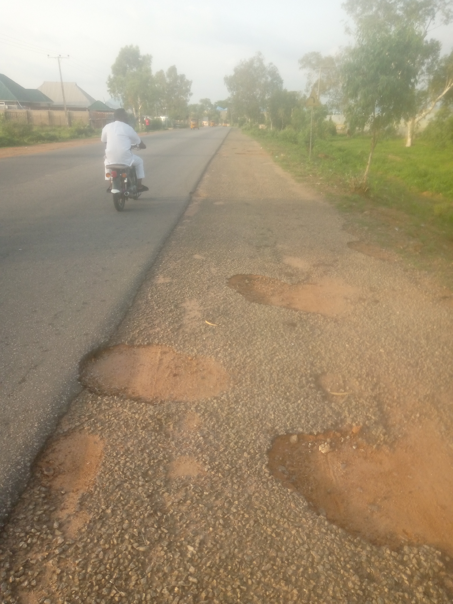 The image depicts a paved road in Jalingo, Nigeria, with a person riding a motorcycle in the left lane, traveling away from the camera. The rider is wearing a white robe. The road surface on the right side of the image is damaged, with several large potholes filled with reddish-brown earth and gravel. Trees line the right side of the road, and in the background, buildings and more trees are visible under a hazy sky. The overall scene suggests a semi-urban or rural environment with infrastructure in need of repair.