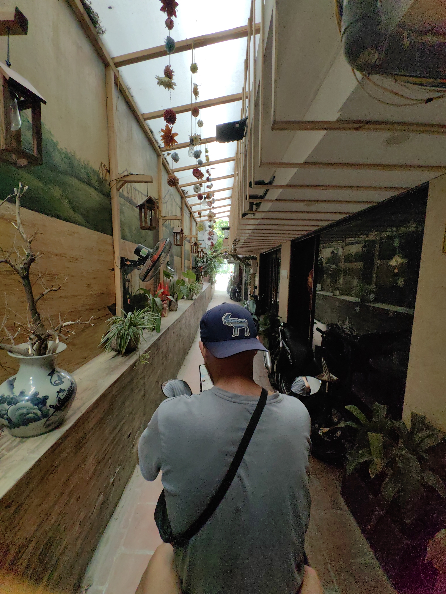 This media file captures a narrow, covered alleyway or passageway in what appears to be Hà Nội, Vietnam, viewed from behind a person on a motorcycle or scooter.
The primary subject is a person seen from the back, wearing a light gray t-shirt, a black shoulder bag, and a blue baseball cap with a white deer or moose emblem. They are positioned as if driving or standing on a motorcycle, which is partially visible beneath them.
The scene is an enclosed, elongated space. To the left, there's a decorative wall featuring a painted mural depicting a green landscape with hills. Along this wall, a raised ledge holds various potted plants, a wall-mounted fan, and a large blue and white ceramic vase containing dried branches. Several wooden, birdhouse-like decorative elements hang from the wall or ceiling structure. The ceiling above is made of translucent panels supported by wooden beams, through which ambient light filters. Numerous colorful, artificial flowers or star-shaped ornaments are suspended from the ceiling.
To the right, the alley is lined with storefronts or residential entrances, with several motorcycles parked along this side. The overall lighting suggests daytime, possibly an overcast day or a heavily shaded interior space. No specific activity beyond the person moving through the alley is clearly depicted. No discernible text is visible in the image.
