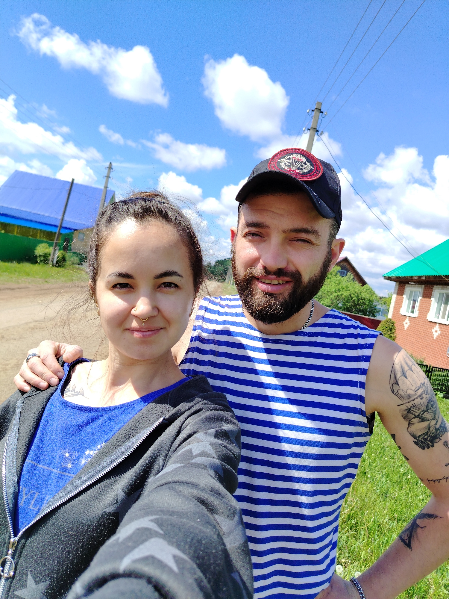 A selfie photograph shows a man and a woman outdoors on a sunny day in Гожан, Russia.
The woman, on the left, has dark hair pulled into a bun, brown eyes, and a nose ring. She wears a blue T-shirt under a black zipper hoodie with star patterns. Her left hand has a silver ring and rests on the man's shoulder. A small tattoo is visible on her chest.
The man, on the right, has a beard and wears a dark baseball cap with a parachute patch. He is wearing a blue and white striped tank top. His left arm is tattooed with a sailing ship, and a silver bracelet is on his wrist.
Behind them, a dirt road runs past houses with blue and green roofs. A utility pole with wires is visible. The sky is blue with scattered white clouds. The background environment indicates they are in a rural or semi-rural area.