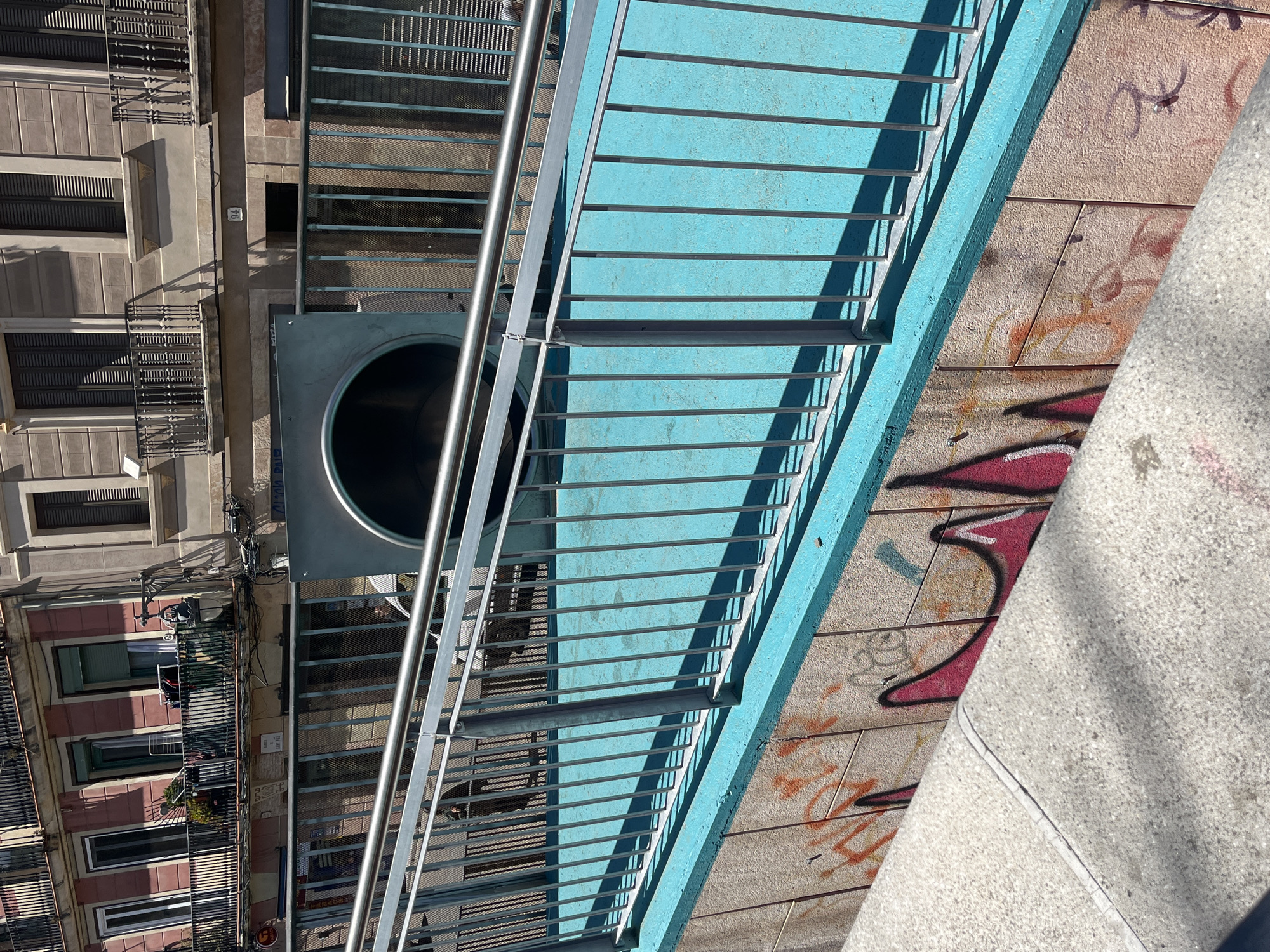 An elevated, bright blue pedestrian walkway or bridge with silver metal railings diagonally crosses the frame, appearing to be suspended above a street or alley. The blue surface shows signs of wear and has a textured finish. Strong, elongated shadows cast by the railings fall across the walkway and the structures below, indicating bright, direct sunlight.
To the left and below the walkway, a multi-story urban building is visible, featuring numerous windows, closed dark shutters, and ornate balconies. The building's facade is primarily light brown with some reddish-brown sections. A large, metallic, cylindrical structure, resembling a slide or chute, is integrated into the walkway's left side, with its circular opening facing downwards.
To the right and below the walkway, a light-colored, textured concrete wall or ground surface is heavily adorned with various graffiti tags and abstract shapes in shades of red, orange, and black.
No people are visible, and no specific activity is taking place. The scene is static, capturing an architectural urban landscape under clear, sunny weather, likely mid-day due to the distinct shadows. The building's style and the presence of graffiti are consistent with a European city environment, supporting the location context of Barcelona, Spain. The visible text consists only of illegible graffiti and very small, indistinct lettering on distant signs.