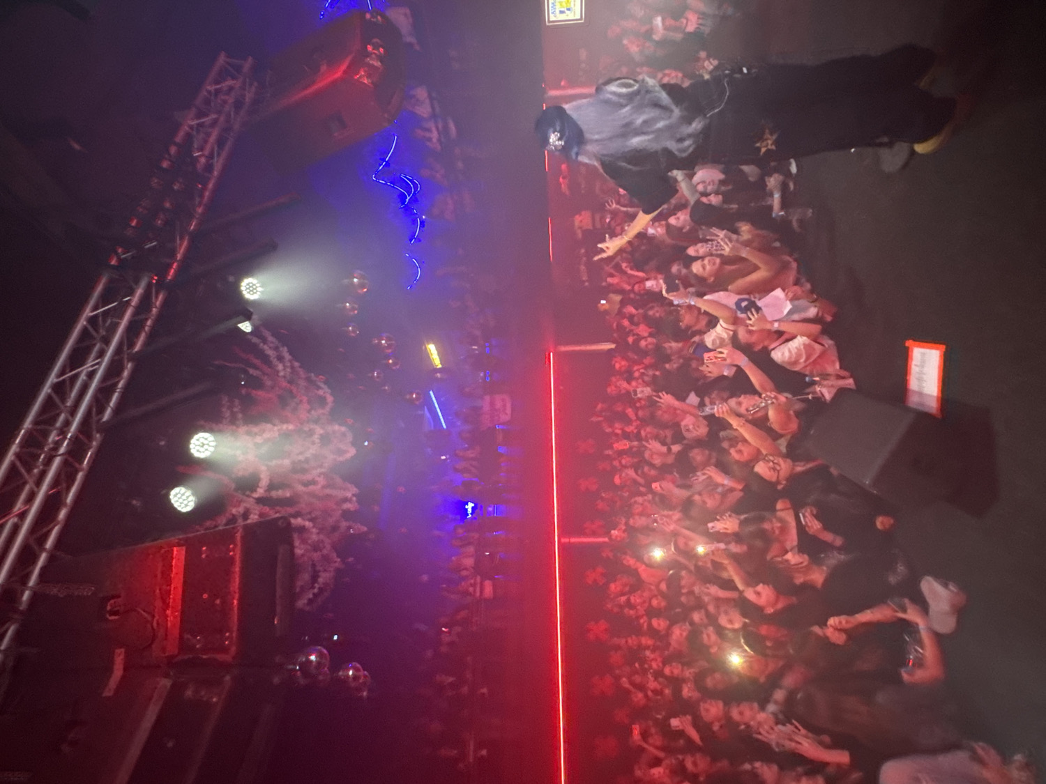 This media file captures a vibrant indoor concert or club event in Vladivostok, Russia, from an elevated perspective looking down and across the venue. The scene is dominated by a large, enthusiastic crowd of young people, densely packed and extending from the bottom to the right side of the frame. Many attendees are holding up cell phones, illuminating their faces and recording the performance.
On an elevated stage or platform towards the center-top of the image, a performer with long, distinctive grey or white hair, dressed in dark attire, is visible. They are actively engaging with the audience, with one arm extended towards the crowd.
The left side of the frame showcases stage infrastructure, including tall metal trussing and large loudspeakers suspended overhead. Bright white spotlights cut through atmospheric haze or smoke, illuminating sections of the audience. The venue is bathed in dynamic red and blue stage lighting, with small disco balls reflecting light. A prominent vertical red light strip runs along the edge of the performance area.
Visible text includes a small, partially obscured yellow sign positioned high above the performer, and a small rectangular red sign with white text placed on the stage floor near a speaker on the right. The overall atmosphere is energetic and dark, characteristic of a nighttime live music event, with the crowd's engagement evident through their raised hands and phone lights.