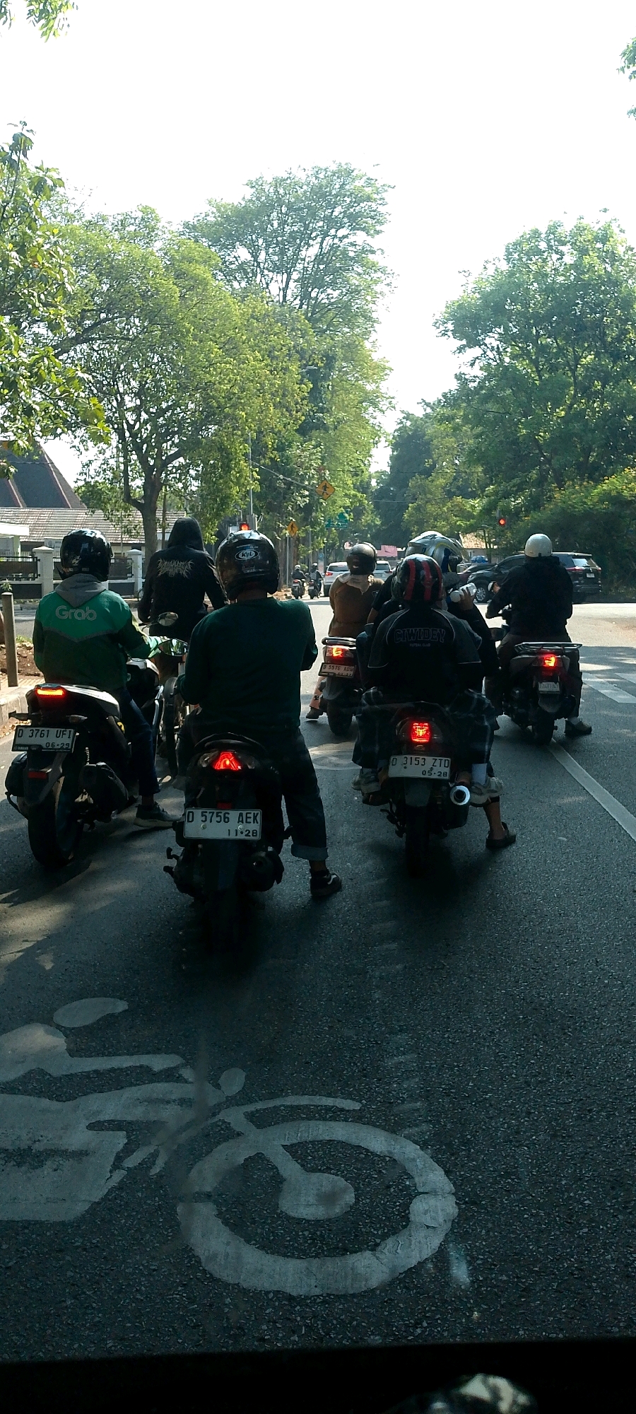 The image captures a traffic scene in Bandung City, Indonesia, likely on a street lined with trees. The perspective is from a vehicle behind a group of motorcyclists, indicating a traffic jam or slow-moving traffic.
In the foreground, several motorcycles are visible from the rear. The closest motorcycle on the left has a rider wearing a green jacket with the "Grab" logo, a ride-hailing service. The rider is wearing a black helmet. The license plate of this motorcycle reads "D 3761 UF1 06-29".
To its right, another motorcycle has a rider wearing a black hoodie with a graphic on the back and a black helmet.
Further back, a rider in a green shirt and black helmet is visible.
The motorcycle to the right of this rider has a rider wearing a brown outfit and a beige helmet. The license plate is partially obscured but appears to start with "D 5576".
In the middle ground, more motorcycles are visible, some with riders and others without clear occupants. The overall scene suggests a typical urban commute.
The environment includes lush green trees lining the road, indicating a well-treed area within the city. The sky is bright, suggesting daytime, likely morning or afternoon, with the sun casting shadows. A white bicycle lane marking is visible on the asphalt in the lower portion of the image, suggesting the presence of a designated cycle path. Traffic signals and street signs are visible in the background, further confirming an urban setting. The road surface appears to be asphalt.