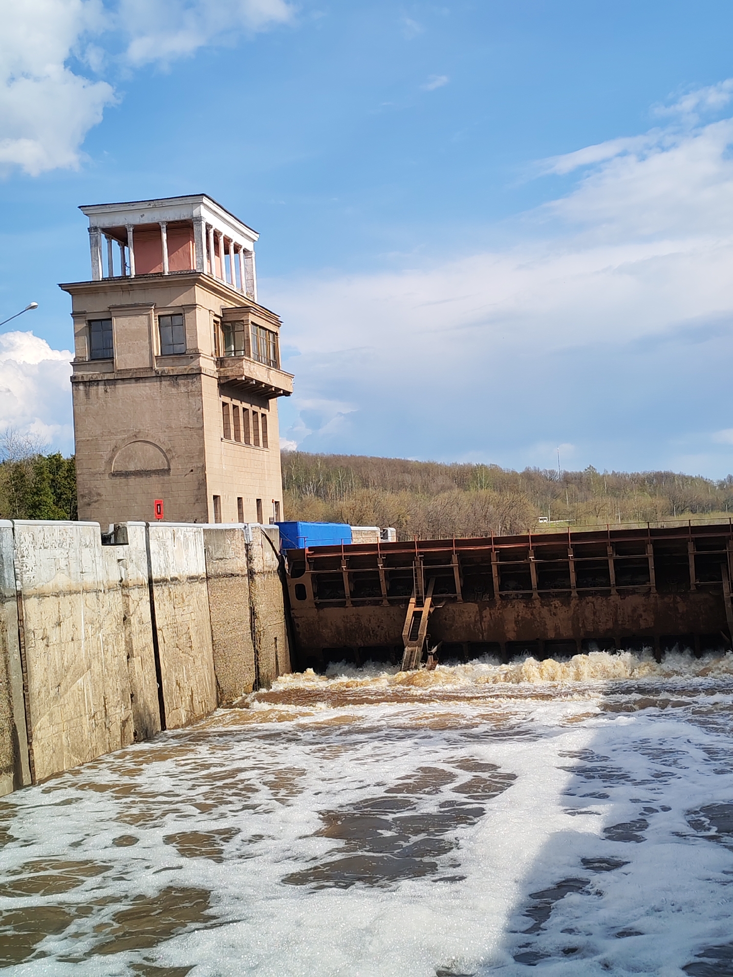 The image shows the Yakhroma lock (number 1) on the Moscow Canal in the city of Yakhroma, Russia. The lock is releasing water, causing frothy waves. The lock's control tower is visible to the left, with a distinct architecture featuring a classical colonnade on top. Behind the lock, there is a forested area. The sky is blue with scattered clouds.