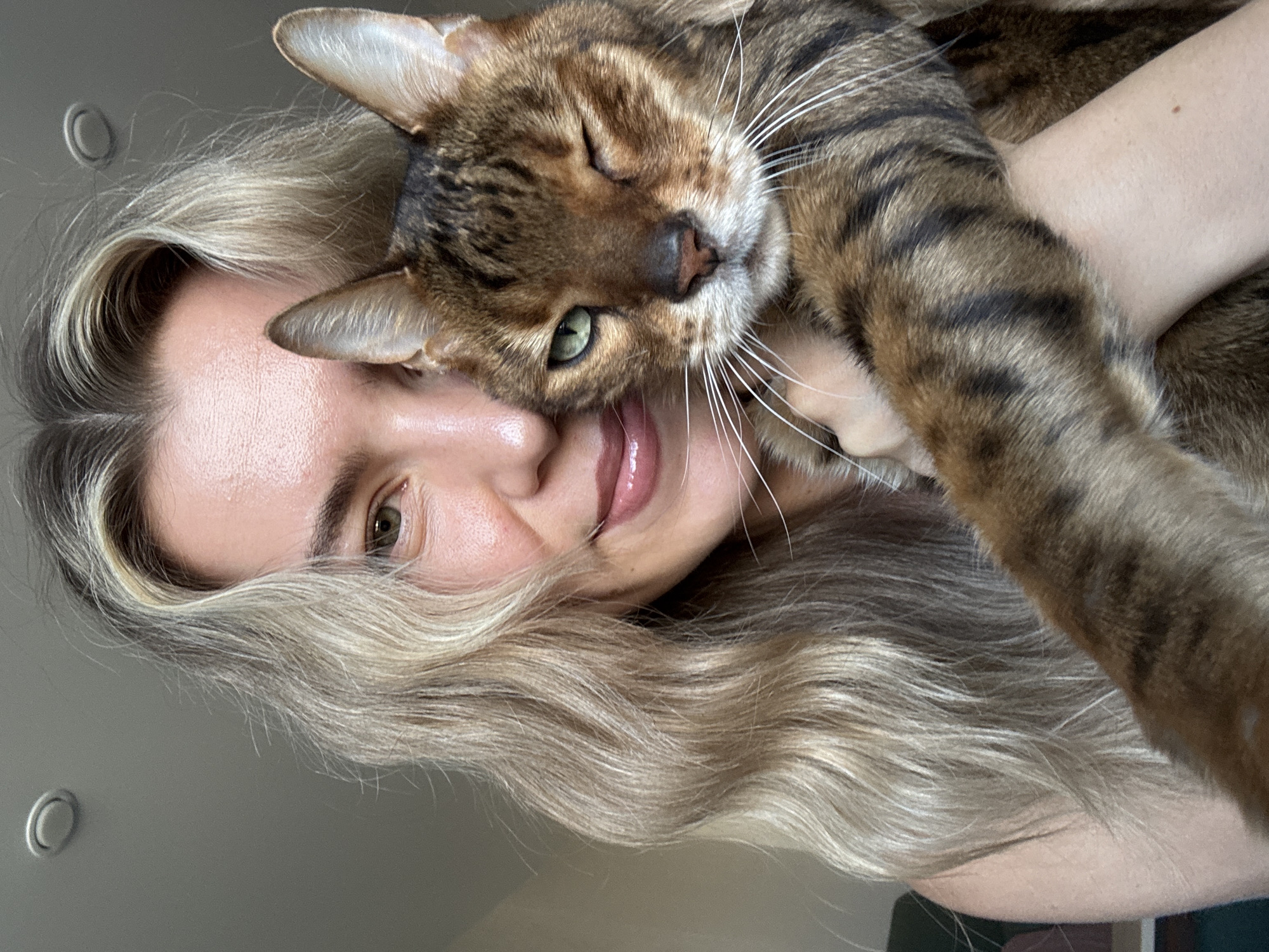 A close-up, rotated portrait captures a person affectionately holding a cat. The person, positioned on the right side of the frame, has long, wavy blonde or light brown hair that cascades around their face. Only a portion of their face is visible, including one eye with a greenish-brown iris, the bridge of their nose, a cheek, and their mouth curved into a gentle smile, with glossy pink lips. Their skin appears fair.
Cuddled close to the person's face is a domestic cat, likely a Bengal, identifiable by its distinct brown and black spotted and striped fur pattern. The cat's head is prominent, with one eye partially closed, giving the impression of a wink, and its bright green iris visible. Its nose is a soft pinkish-brown, and it sports long, prominent white whiskers. The cat's body and paws are draped over the person's arm and shoulder, suggesting a comfortable and close embrace.
The activity depicted is one of shared affection between the person and their pet. The cat appears relaxed and content, possibly nuzzling, while the person's smile conveys happiness and fondness for their feline companion. The background is an out-of-focus, plain light grey or off-white interior wall, with a subtle circular light fixture or hook visible in the upper left corner of the frame. The soft, even lighting suggests it is daytime, possibly indoors near a natural light source. There is no visible text in the image.