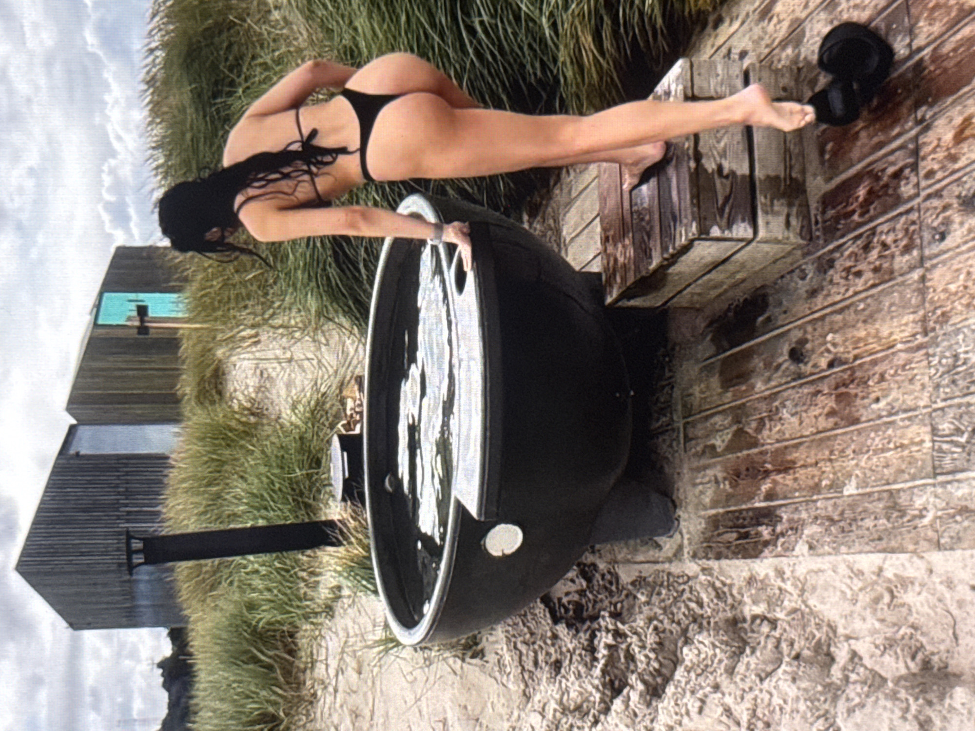 A woman in a black bikini is leaning over a black hot tub filled with water. She is positioned with her back to the camera, her legs extended and her hands on the rim of the tub. The hot tub is situated outdoors on a wooden deck, with sand and dry grass surrounding it. In the background, there are two corrugated metal structures and a cloudy sky. The scene appears to be in a coastal or beach area, given the sand and vegetation. The lighting suggests it could be daytime, with overcast conditions. No text is visible in the image.