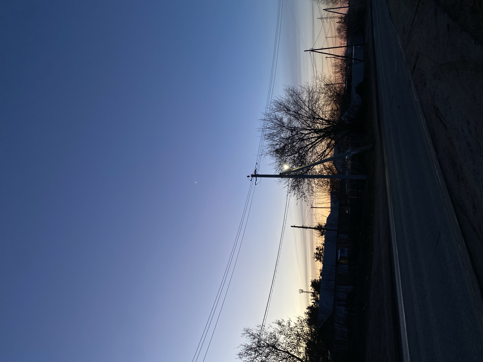 The image shows a dusk or dawn scene in Chaikovsky, Russia. The sky transitions from a deep blue to a light gradient with hints of orange near the horizon. A crescent moon is visible in the dark blue part of the sky. In the foreground, there is a road running along the right side of the frame. Along the roadside, there are bare trees and utility poles with power lines stretching across the sky. Beyond the trees, there appear to be buildings and potentially other vegetation silhouetted against the horizon. The overall scene suggests a rural or suburban setting.