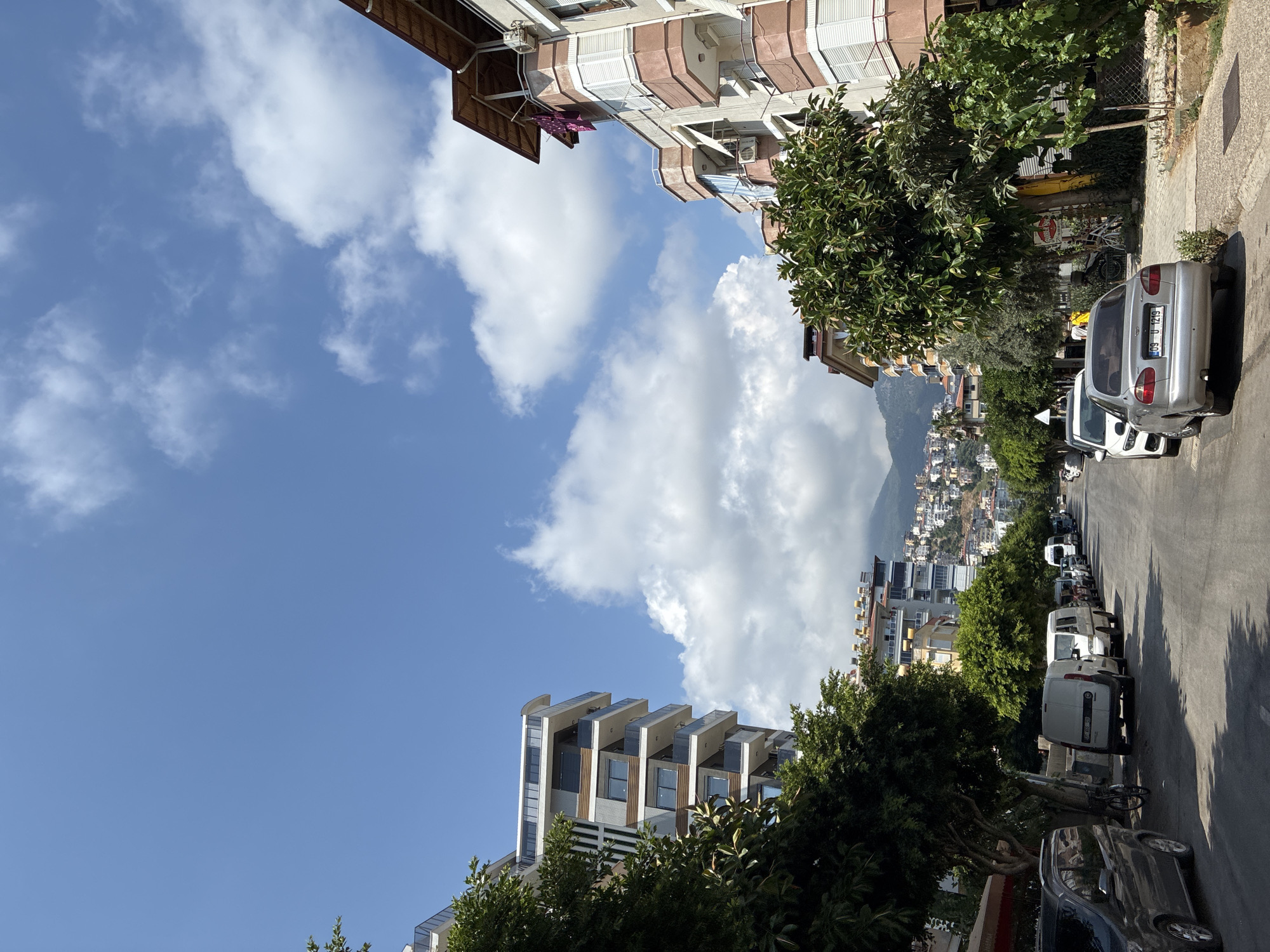 The image depicts a street scene in Alanya, Turkey, on a clear day with scattered clouds. The street is lined with buildings and trees, with cars parked on either side. The foreground features a silver sedan parked on the right side of the road. The street extends into the distance, revealing more buildings and a hillside covered in structures, indicating an urban environment on a slope. The sky is a vibrant blue, with fluffy white clouds adding texture. The architecture of the buildings suggests a Mediterranean style. The overall impression is one of a lively, sunny day in a coastal Turkish city.