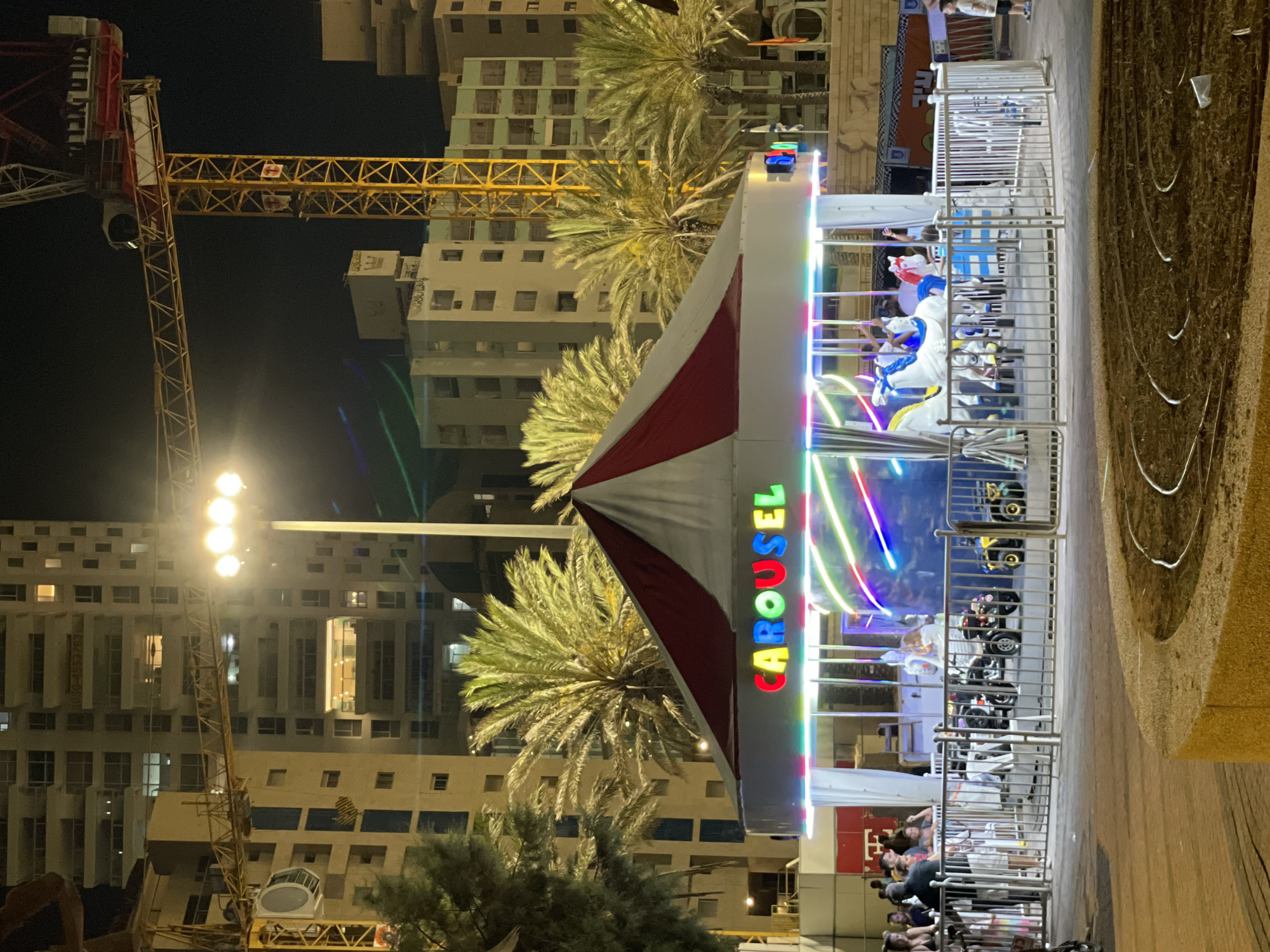 The image shows a carousel with the word "CAROUSEL" written in colorful lights. The carousel has white horses and other rides. The carousel is surrounded by a metal fence. People are standing around the carousel, some are watching. The carousel is lit up with colorful lights. In the background, there are tall buildings and palm trees. There are also cranes in the background, suggesting construction. The overall scene appears to be a nighttime view of a recreational area in the city of Netanya, Israel. There is a paved area in the foreground, with some brown dirt and circular patterns. The image has a slightly grainy quality, consistent with low-light photography.