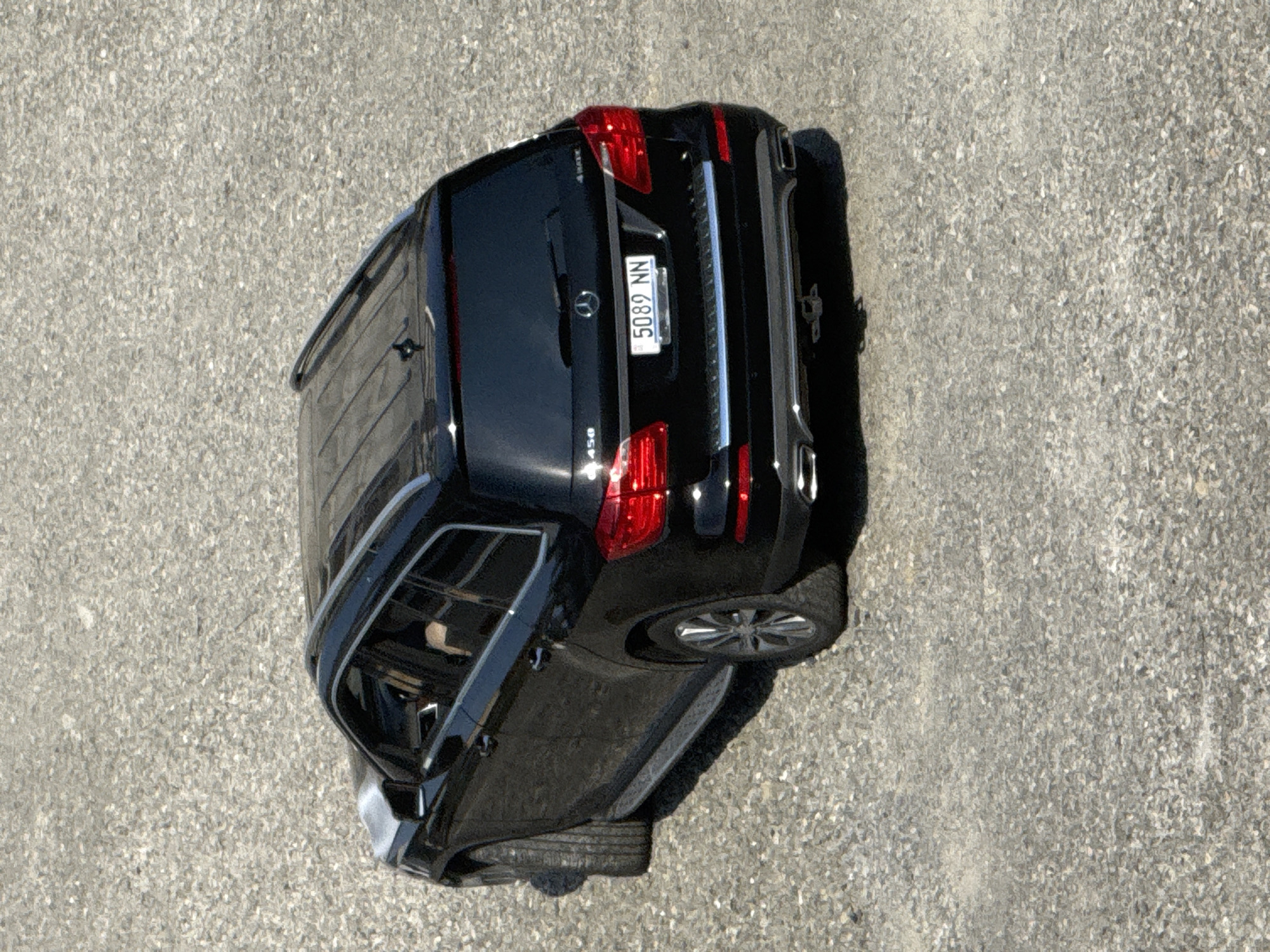 This is a top-down, slightly angled aerial view of a black Mercedes-Benz GLS SUV parked on a coarse asphalt surface. The vehicle is the primary subject. The scene appears to be outdoors during daylight, with no discernible weather conditions mentioned. The ground texture is consistent with a road or parking lot.
Notable details include the car's license plate, which clearly displays the text "5089 NN". The car's rear is prominently featured, showing its taillights, exhaust pipes, and a part of the trunk. The overall impression is of a static scene, with the car simply parked. There are no people visible in the image. The location context is given as Tbilisi, Georgia.