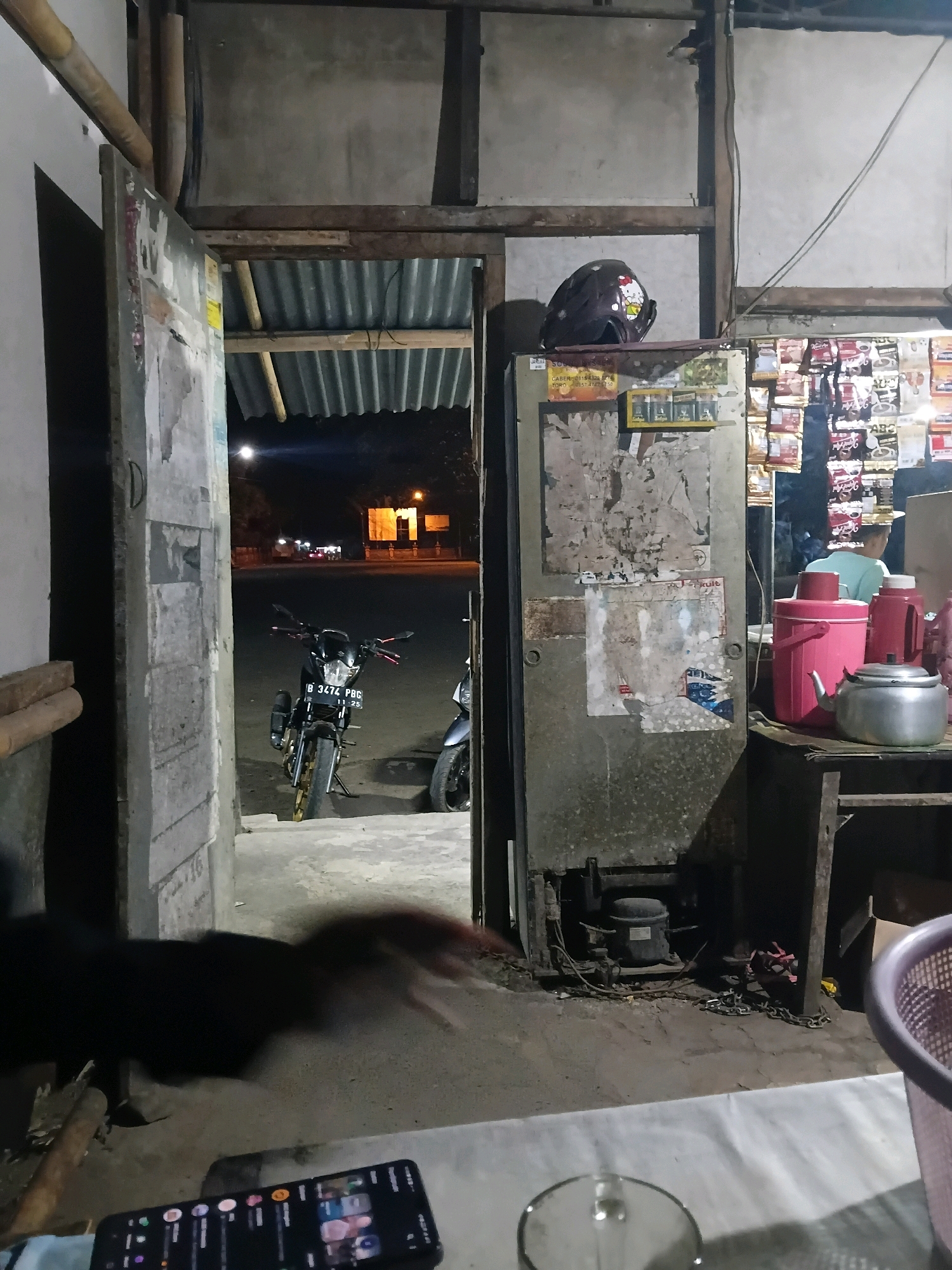 The image captures a dimly lit, cluttered storefront or workshop at night in Bantarbarang, Indonesia. A doorway on the left reveals a street scene with two motorcycles parked outside, one in the foreground with a license plate reading "B 3474 PBG". The interior is filled with various items: a dark-colored helmet with a character design sits atop a refrigerator, which is covered in peeling posters and flyers. To the right, shelves are stocked with numerous small packages of what appear to be food items or snacks. A person in a light-colored shirt is partially visible behind the counter, looking towards the shelves. On a wooden surface in the foreground, there are two large thermal beverage containers, one pink and one white, along with a silver teapot. The floor is concrete, and a dark, blurry object, possibly a dog or a person in dark clothing, is in motion in the lower left corner. A smartphone with its screen illuminated is also visible on a white surface in the lower foreground. The overall environment suggests a small, informal business operating in a rural or semi-urban setting.