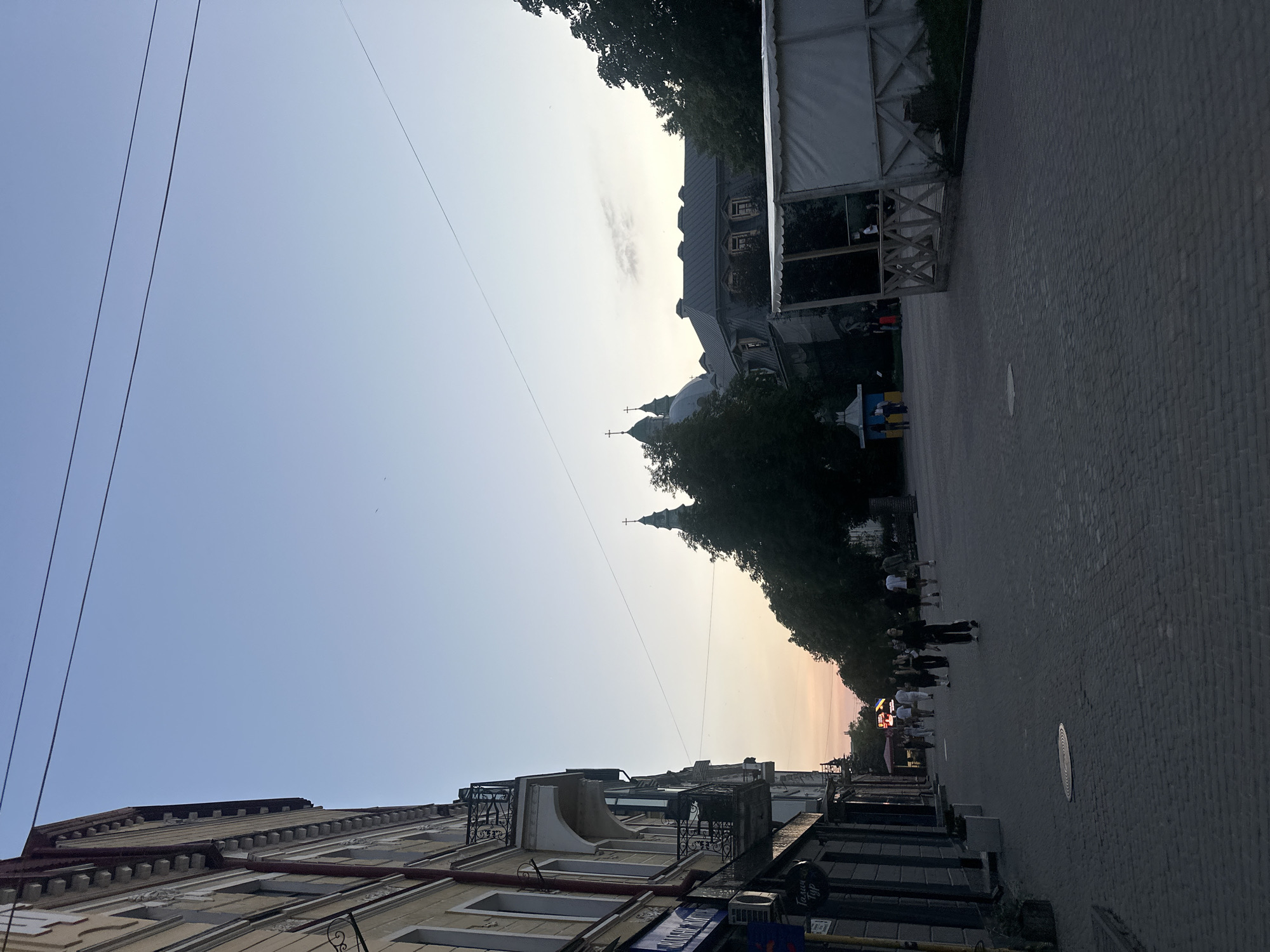 The image shows a street scene in Ternopil, Ukraine, likely during the late afternoon or early evening, given the soft lighting and pale sky. On the left, a multi-story building with ornate balconies runs along the street. The building has light-colored facades and dark windows. Several shops are visible on the ground floor, with signage that is partially obscured. A cobblestone sidewalk and street dominate the foreground.
In the distance, several people are walking along the street, some individually and some in small groups. They are dressed in casual clothing. Beyond the people, trees line the street, partially obscuring a large building with a grey, pitched roof and multiple windows. In the background, two green domes topped with crosses suggest a church or cathedral. A white tent-like structure with wooden framing is visible on the right side of the frame, suggesting a temporary setup or market stall. Overhead, power lines are visible against the pale sky. The overall atmosphere appears to be relaxed and urban.