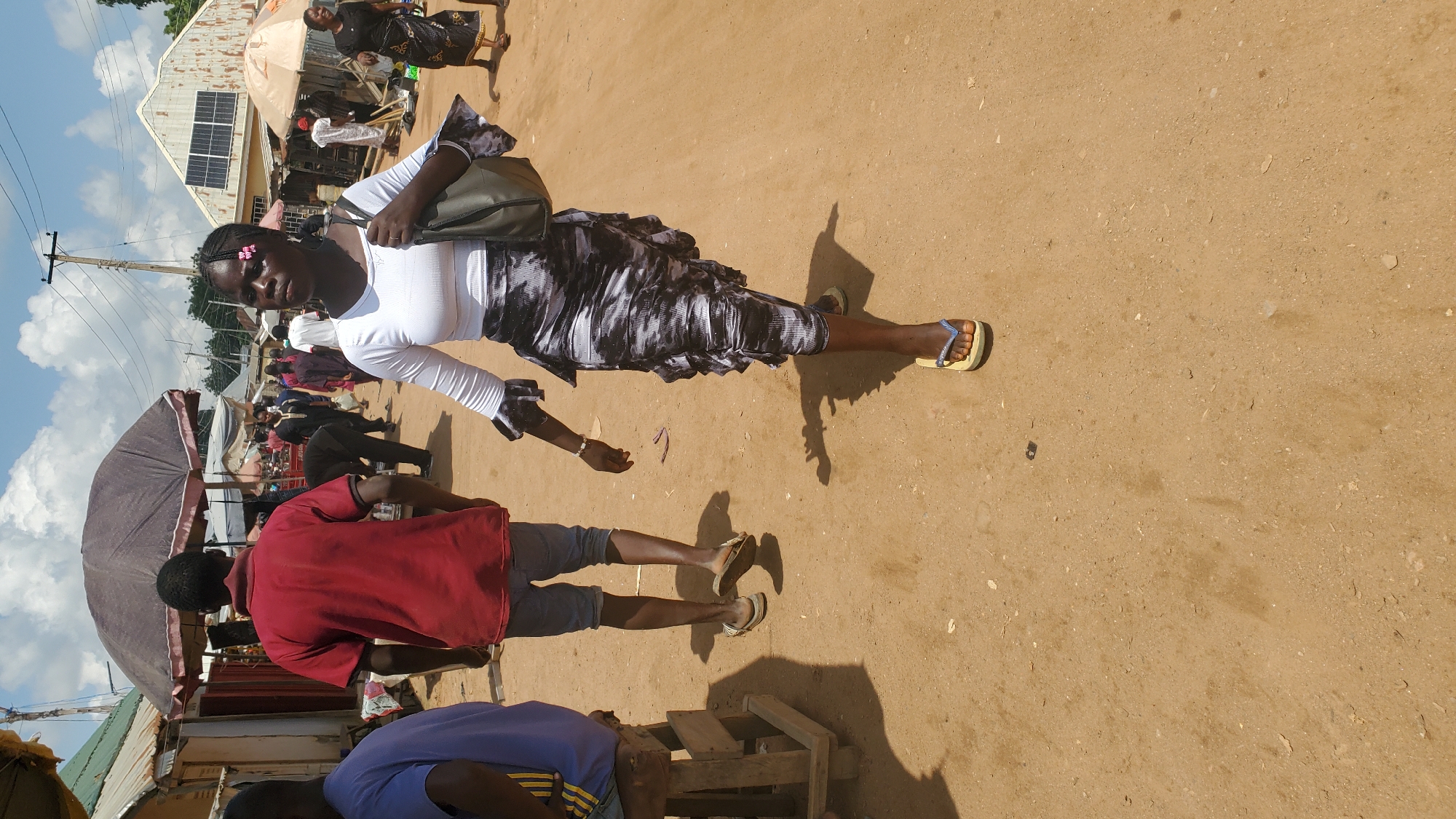 In Jalingo, Nigeria, a woman walks forward on a dirt path with a textured, patterned skirt and a white, long-sleeved top. She carries a dark-colored shoulder bag and wears light-colored flip-flops. Behind her, a man wearing a red shirt and denim shorts walks away from the camera. To the left, a person in a blue t-shirt sits on a wooden stool. The scene is outdoors with stalls and market activity in the background under a partly cloudy sky.