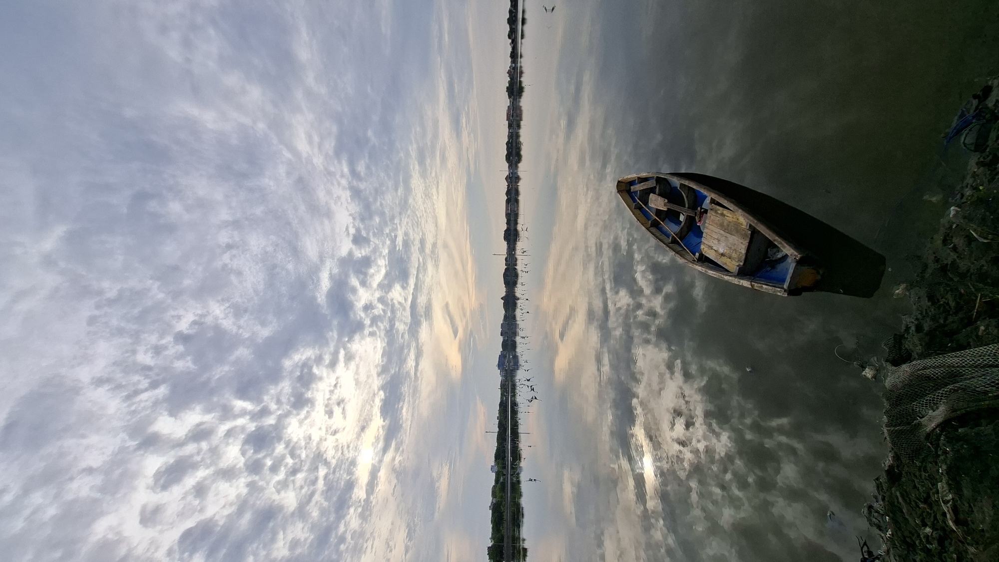 The image depicts a serene waterscape at dawn, likely in Lhokseumawe, Indonesia. A small, weathered wooden boat with blue trim floats gently on the calm water, its reflection a near-perfect mirror image. The boat contains a simple wooden bench and a paddle. In the distance, a line of buildings and structures forms the horizon, softened by the morning mist. A flock of birds, appearing as small white specks, are scattered across the water in the mid-ground. The sky is filled with a dramatic display of clouds, streaked with the pale hues of sunrise, creating a textured ceiling above the scene. The water's surface is highly reflective, capturing the intricate patterns of the clouds and the distant shoreline. The foreground on the left shows a muddy bank with some discarded fishing nets and debris, indicating a coastal or riverine environment. The overall atmosphere is tranquil and contemplative, capturing a quiet moment before the day fully begins.