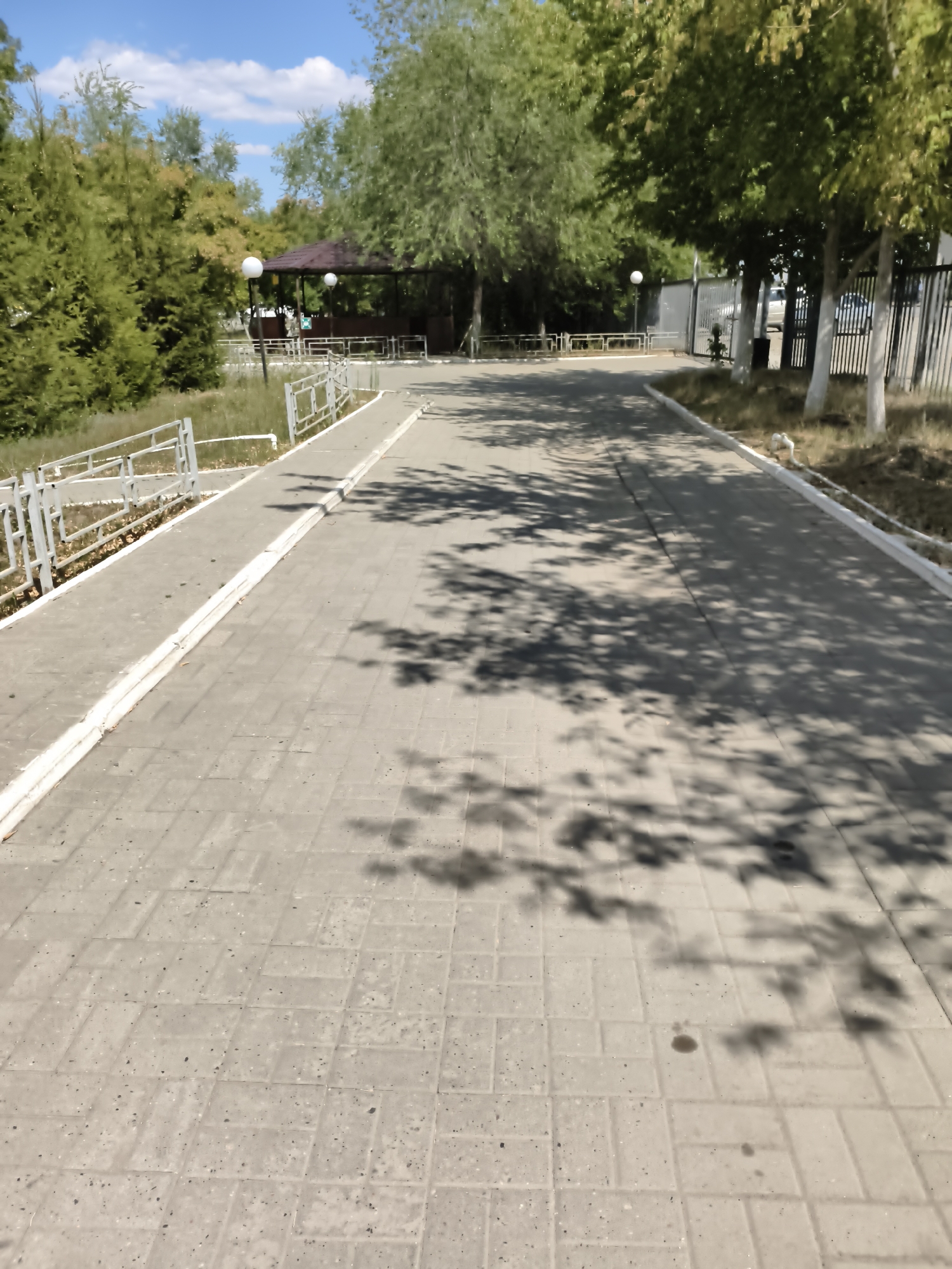 The image shows a paved walkway in a park-like setting. The walkway is made of light-colored rectangular pavers and is bordered by a low white-painted curb. To the left of the walkway is a grassy area with a white metal fence and some trees and bushes. In the distance, there is a brown gazebo with white lights. To the right of the walkway, there is a black metal fence, some trees with white trunks, and dry grass. The ground is covered with large, distinct tree shadows, suggesting it is daytime with bright sunlight. The sky is blue with scattered white clouds. The location is identified as a park area in the city of Aktobe, Kazakhstan.