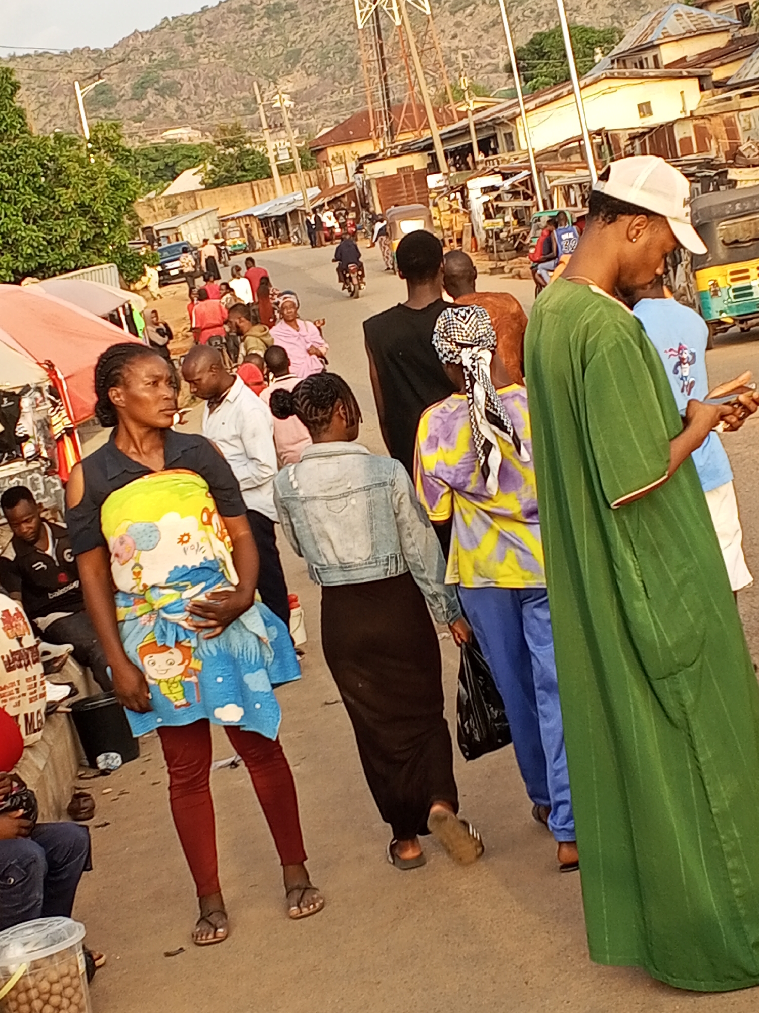 The image depicts a street scene in Gwong, Nigeria. A group of people are walking on a paved road. In the foreground, a woman wearing red leggings, sandals, and a black shirt with a patterned cloth wrapped around her is walking. To her right, another woman wearing a denim jacket and a long black skirt is also walking. Further to the right, another woman is wearing a colorful yellow and purple shirt, blue pants, and a patterned headscarf. To the far right, a man is wearing a long green garment and a baseball cap, using a smartphone. In the background, there are other people, motorcycles, a tricycle, buildings, and utility poles. In the distance, a hill or mountain is visible. The scene suggests a typical day in the city with people going about their daily activities.