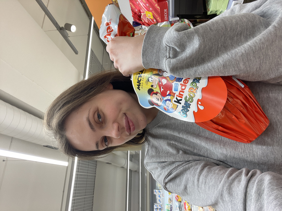 An adult female is depicted in a retail setting, specifically a supermarket aisle in Monsummano Terme, Italy. She possesses light brown hair and a fair complexion, and is wearing a plain grey crew-neck sweatshirt. Her left arm is raised, holding a large Kinder Gran Sorpresa chocolate egg. The egg is enveloped in red foil and features a white and blue cardboard sleeve displaying "Disney Mickey and Friends" characters (Mickey Mouse, Goofy, Donald Duck) along with the brand name. The female is looking directly at the camera with a faint smile. The background consists of white ceiling panels with linear fluorescent lighting fixtures and blurred shelving units stocked with various products, including confectionery like Lindt, indicating a commercial grocery environment.