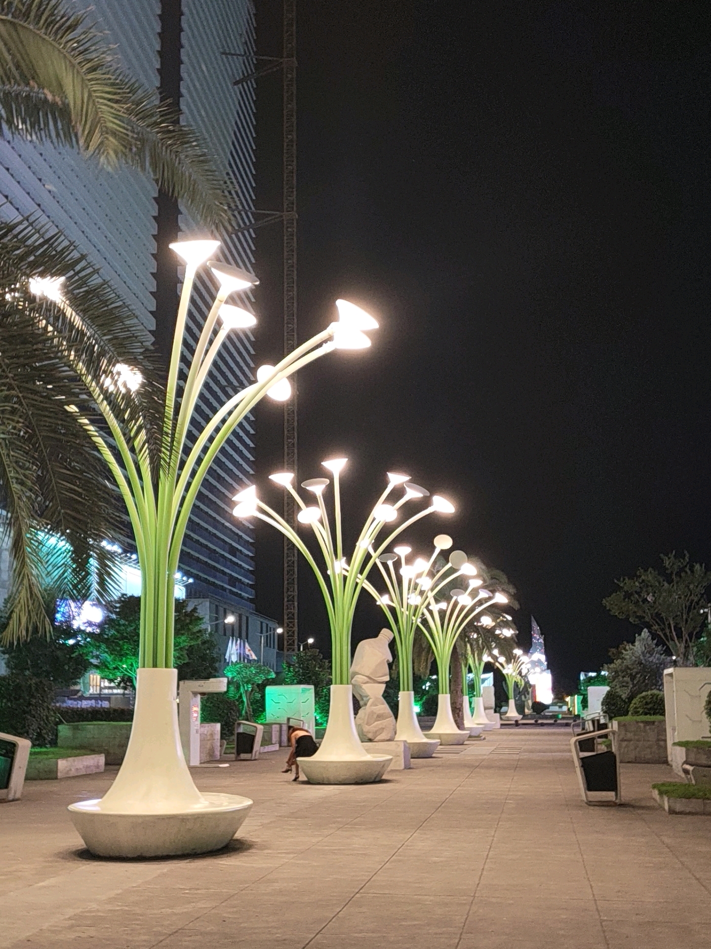 In Batumi, Georgia, at night, a walkway is illuminated by decorative, tall green lights shaped like plants with glowing white bell-shaped flowers. A palm tree on the left partially obscures a modern, striped building. In the middle ground, a woman in a black dress is bending over near a white bench. Several other identical plant-like light fixtures line the walkway, leading towards a distant, illuminated building. Trash receptacles and planters with small green plants are also visible along the path.