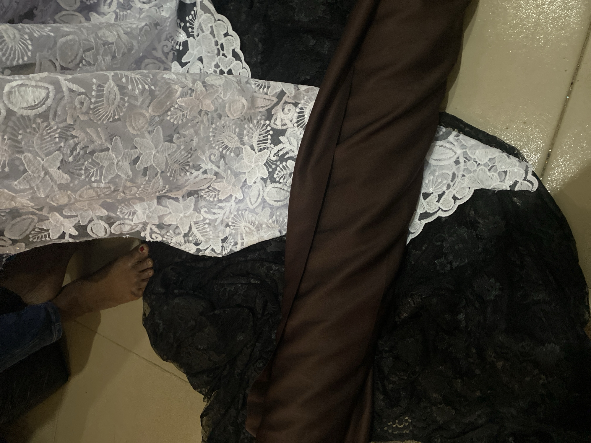 The image shows a pile of fabrics, including white lace and dark brown satin. In the lower left corner, a person's bare foot with red nail polish is visible, with blue denim pants also present. The fabrics appear to be laid out on a textured, light-colored floor, possibly tiled. The overall scene suggests an indoor setting, perhaps a room where fabrics are stored or displayed. The lighting is dim, and there are no clear indicators of the time of day or weather. The focus is on the textures and colors of the fabrics, with the human element providing a sense of scale. The location context is Jalingo, Nigeria, but this information is not visually discernible from the image itself. There is no visible text.