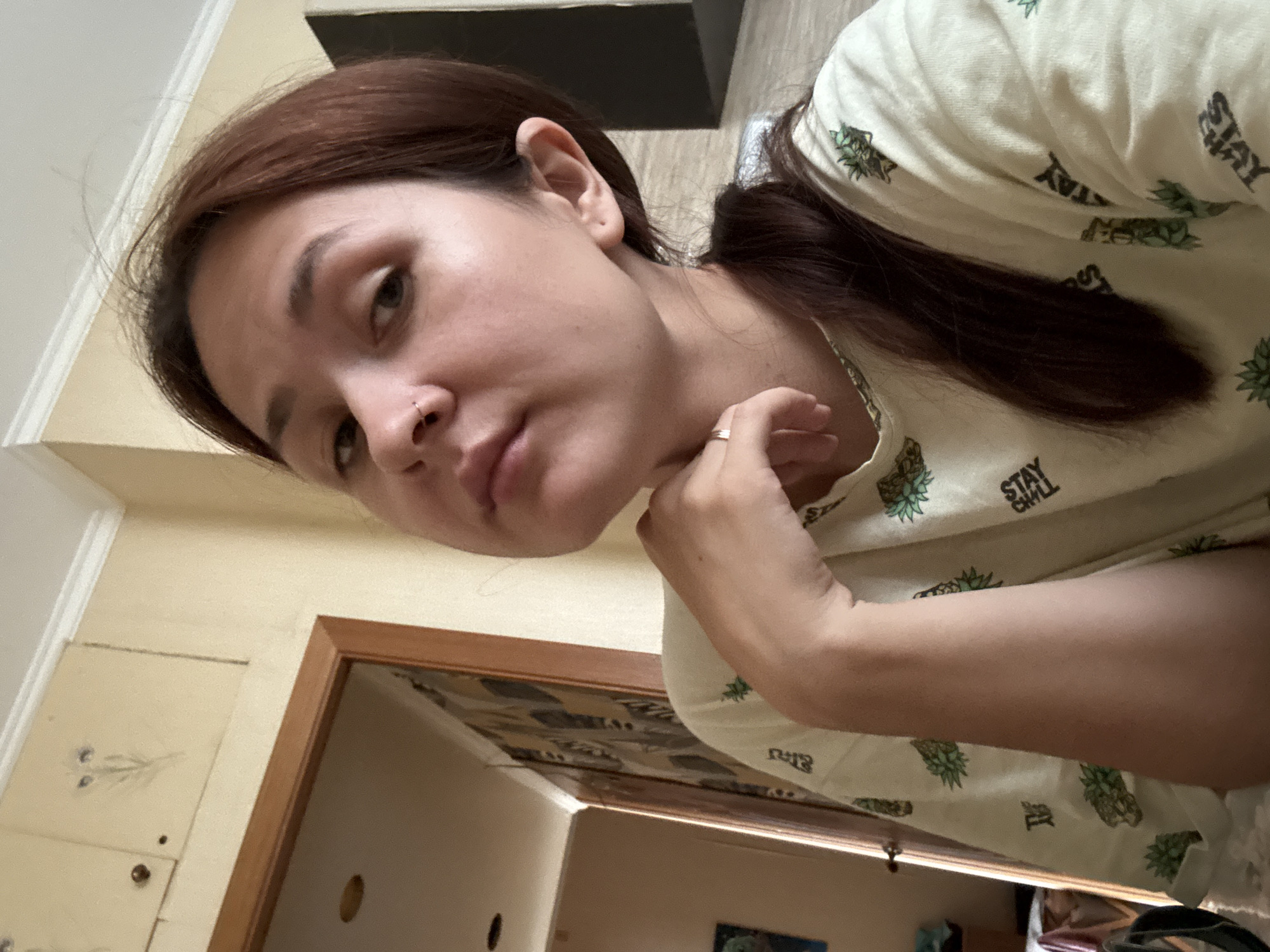 A young woman with brown hair pulled into a ponytail is taking a selfie. She is wearing a light-colored t-shirt with a pineapple pattern and the words "STAY CHILL" printed on it. Her right hand is resting on her chin. The background appears to be an indoor setting, possibly a home, with a doorway and a wall with a patterned wallpaper. The image is taken from a close-up perspective, focusing on the woman's face and upper body. There is no discernible information about the location being in Naberezhnye Chelny, Russia.