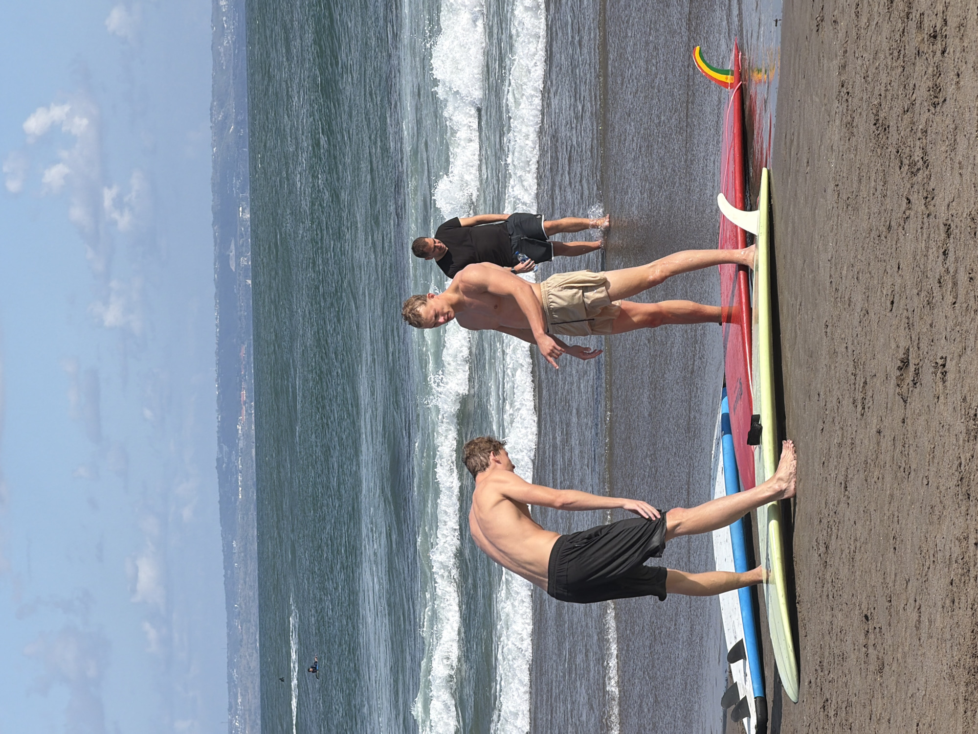 In North Kuta, Indonesia, three men are on the beach with surfboards near the ocean. The man in the foreground is shirtless and wearing black shorts, facing away from the camera and bending down. To his right, another shirtless man in beige shorts is looking forward and gesturing. In the background, a third man in a black t-shirt and dark shorts stands in the shallow water. Several surfboards are on the sand and in the water. The ocean has moderate waves, and a distant coastline with buildings and hills is visible under a partly cloudy sky.