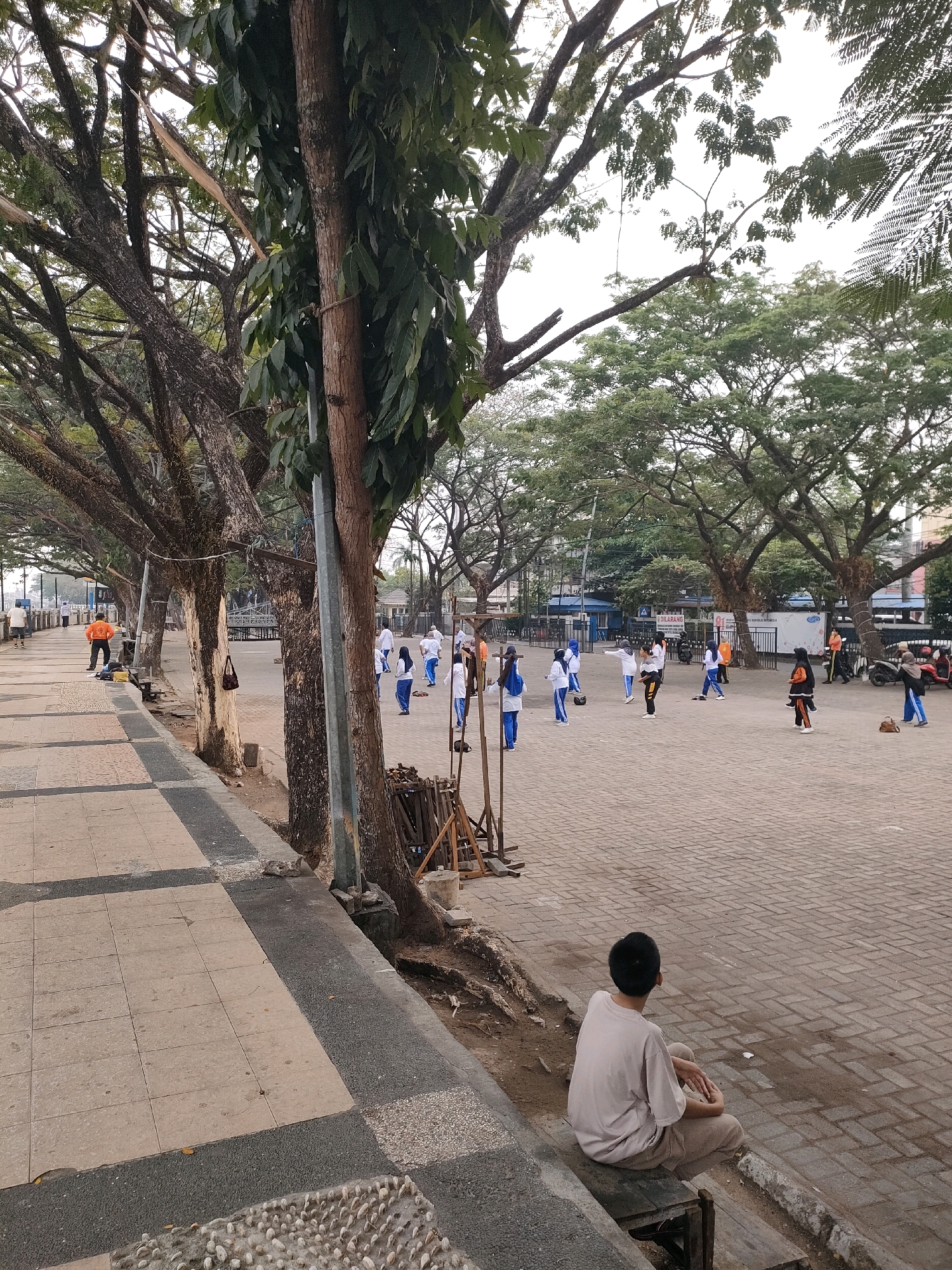 This is an outdoor scene in Banjarmasin, Indonesia, likely a public park or recreational area. In the foreground, a young person with dark hair is sitting on a wooden bench, facing away from the camera, looking towards a group of people in the middle ground. They are wearing a light-colored t-shirt and light-colored pants.
The middle ground features a paved area where several individuals are engaged in what appears to be an exercise or activity. Most of them are wearing white tops and blue bottoms, suggesting a coordinated group. There are also trees with large canopies lining the paved area, providing shade. In the background, buildings and more trees are visible.
The foreground also shows a tiled walkway with a decorative border of small stones. The overall lighting suggests it is daytime, possibly morning or late afternoon, with a slightly overcast sky.
